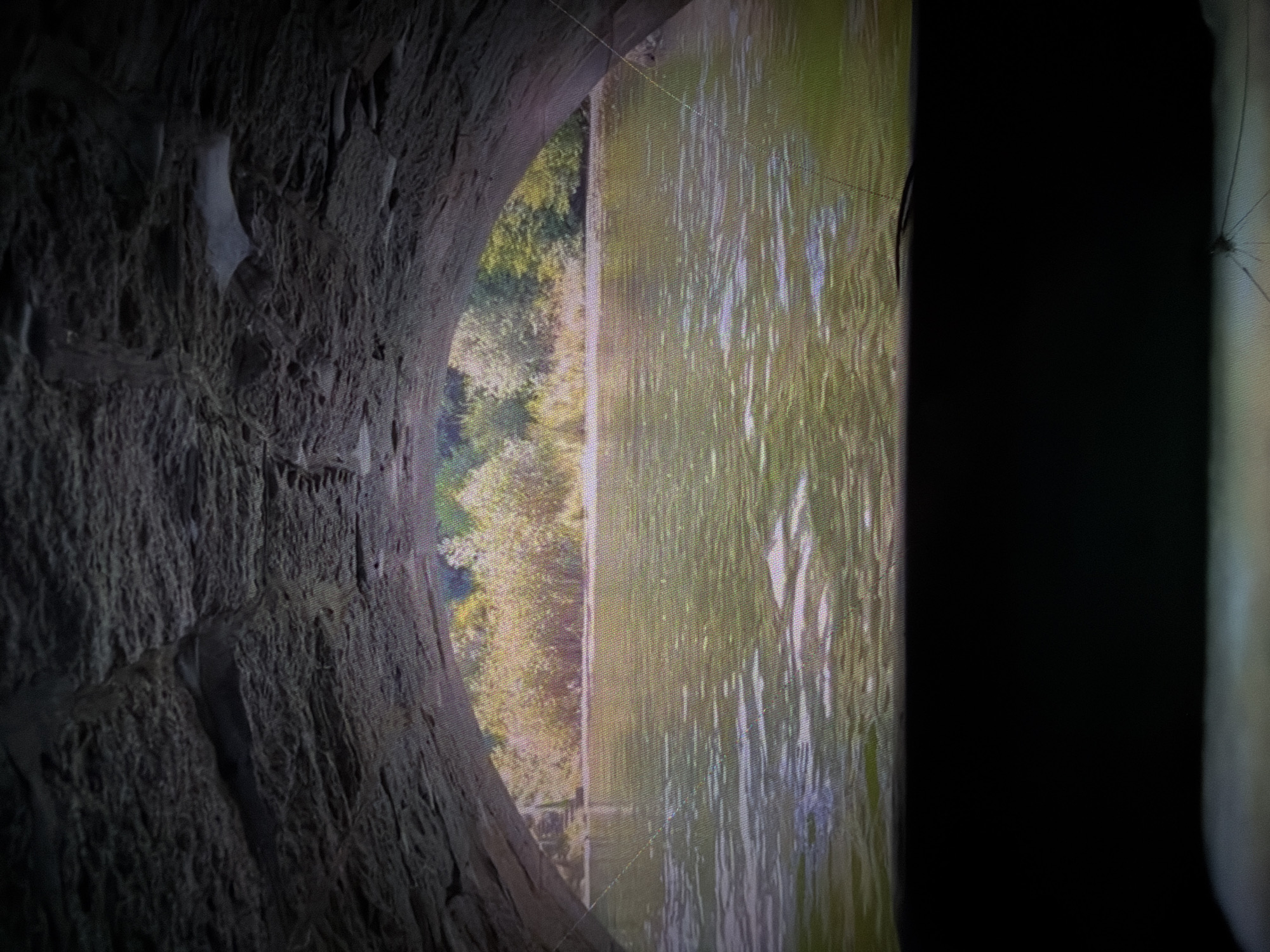 An arched stone structure frames a view of a body of water and a tree-lined shore. The foreground is dark, likely indicating the interior of the archway or a shadowed area. The water in the mid-ground appears rippled and reflects the light. Beyond the water, a dense line of trees with yellow and green foliage forms the background. The sky is not visible. The scene is devoid of any human presence or activity. The location is identified as Unknown, Unknown.