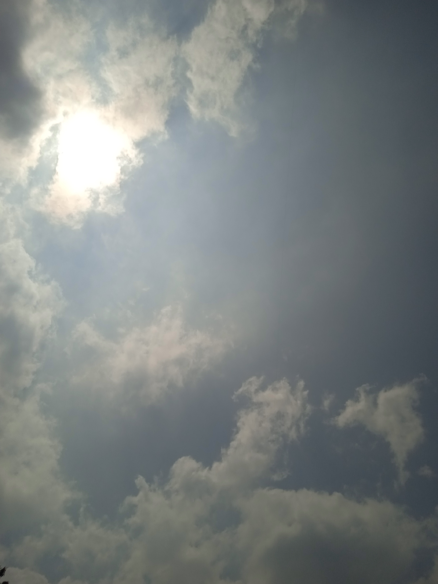 The image shows a view of the sky with clouds and the sun. The sun is partially obscured by clouds, creating a bright, hazy effect. The sky is a mix of light and dark blues and grays, with white and gray clouds scattered throughout. The image appears to be taken from a low angle, looking upwards. The environment suggests an outdoor setting, possibly during the daytime. There are no people or specific man-made objects clearly visible, though the faint presence of what might be foliage or a structure is visible in the bottom left corner. The image quality is moderate, with some noise and a slight blur in certain areas. It is impossible to determine the exact location within Cileunyi, Indonesia, based solely on this image.