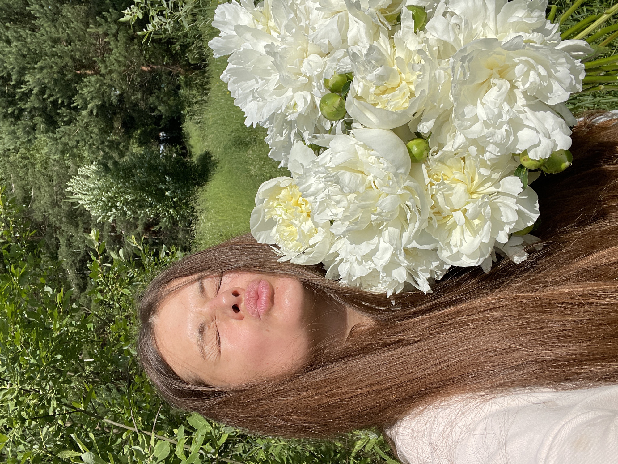 The media file features a young woman lying on her back in a grassy area, with a bouquet of white peonies near her head. She has long, brown hair that is spread around her. Her eyes are closed, and her lips are pursed in a kiss-like gesture. The lighting suggests it is daytime, likely a sunny day, given the shadows cast by the flowers. The background consists of lush green trees and grass, indicating an outdoor setting, possibly a garden or park.
The prominent objects are the white peonies, some in full bloom and others as green buds, adding a delicate and floral element to the scene. The woman's facial expression and the presence of the flowers convey a sense of peacefulness, joy, or playful affection. The overall impression is one of a serene moment captured in nature, with the woman enjoying the beauty of the flowers. The location context provided indicates this scene is in Заречный, Russia. There is no visible text in the image.