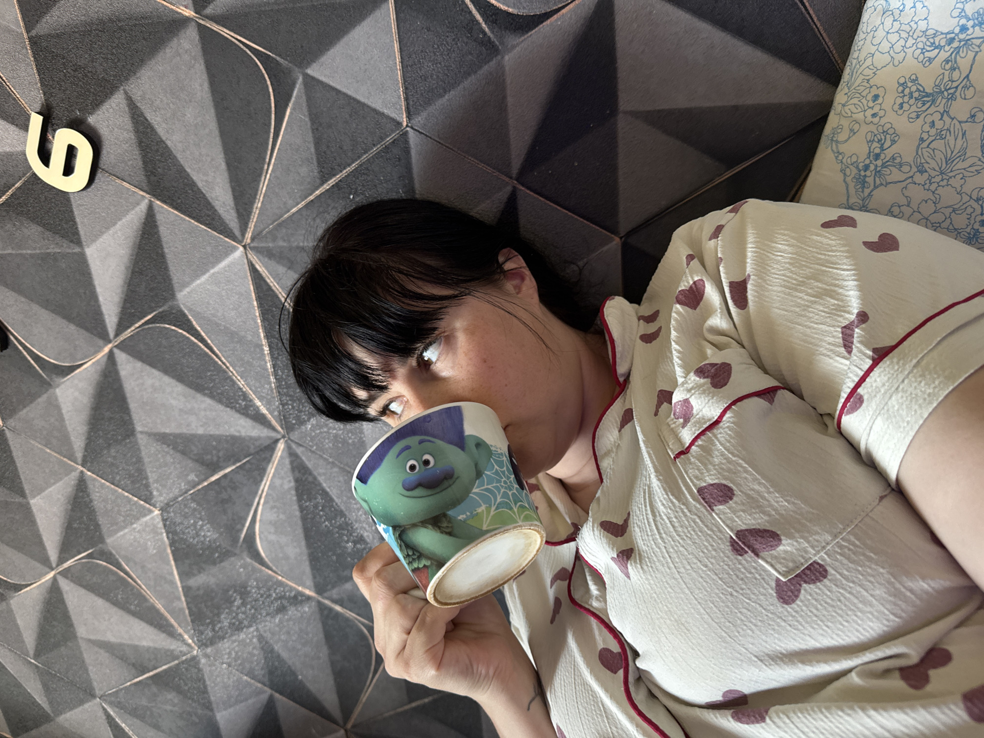 The image shows a woman lying on a bed in a room with a geometric patterned headboard. She is wearing white pajamas with red trim and heart patterns. She is holding a mug with a cartoon troll character on it, and appears to be drinking from it. A number "6" is affixed to the wall. The image location is not discernible as Zav'yalovo, Russia.