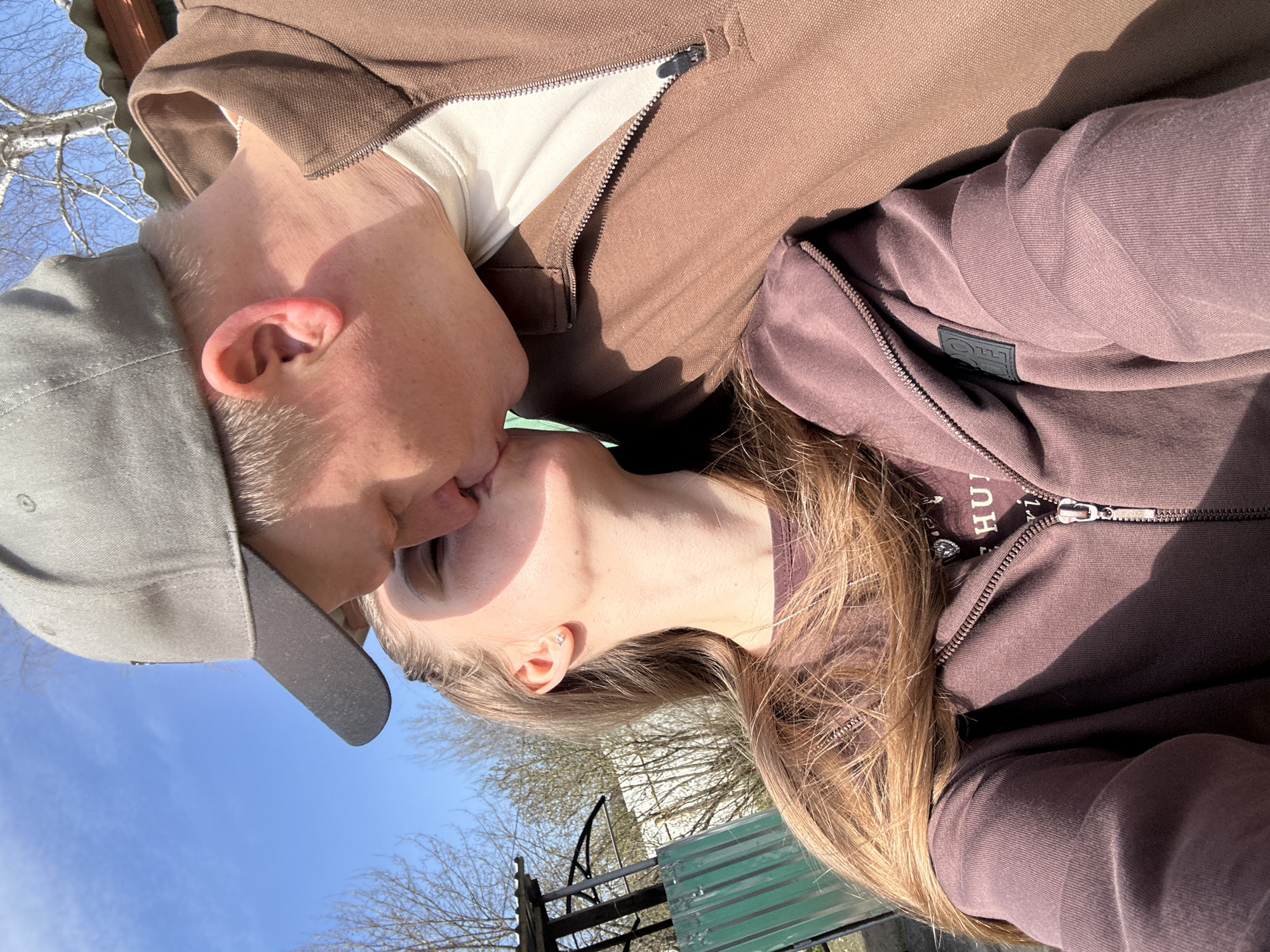 A couple is kissing outdoors on a sunny day. The person on the left is wearing a grey baseball cap and a brown zip-up hoodie. The person on the right is wearing a brown zip-up hoodie, and has blonde hair. The background shows a blue sky, trees, and a green-painted metal structure possibly part of a park bench. The location is unidentifiable as Мещёры, Russia without additional visual clues.