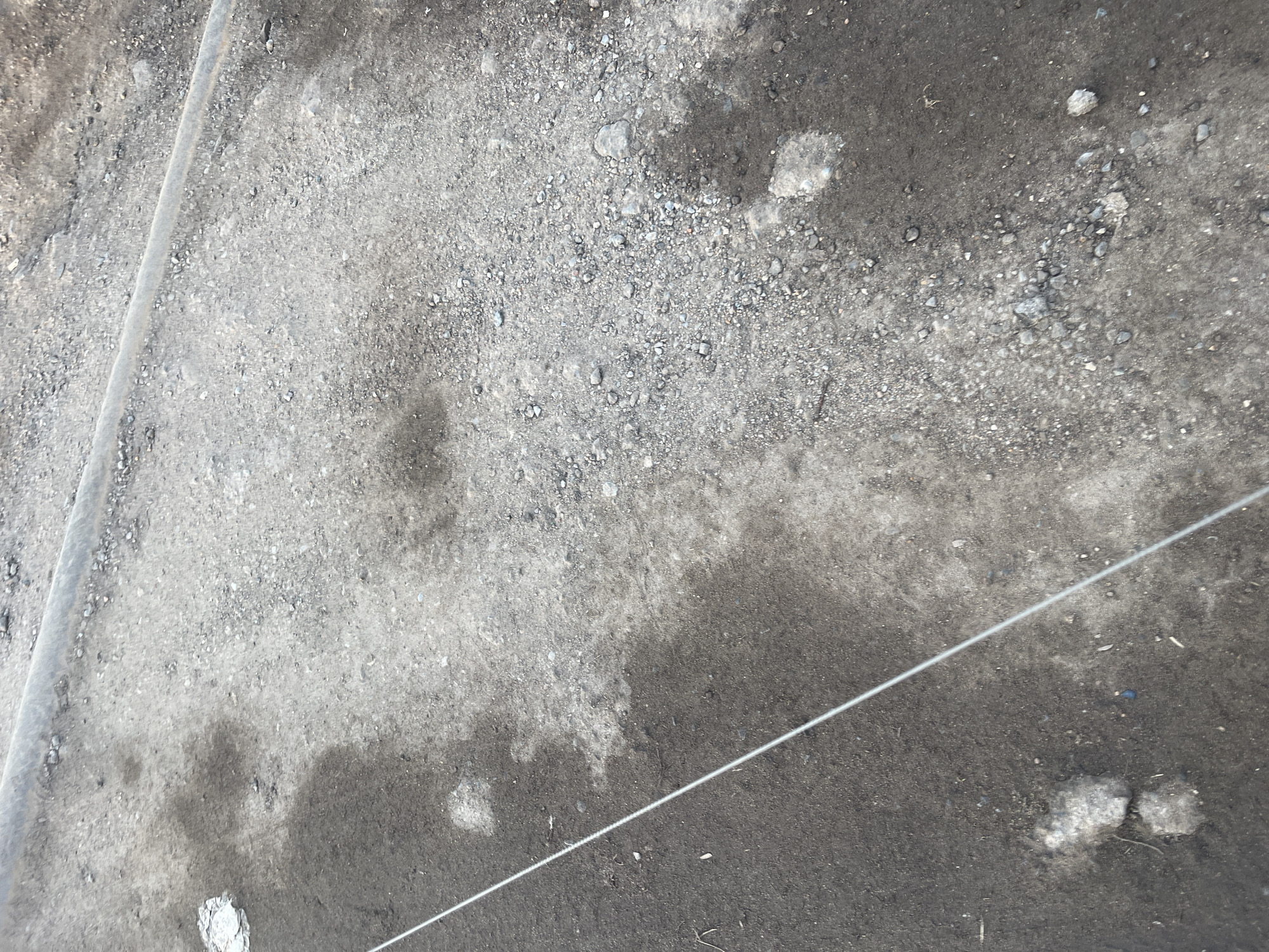 The image shows a section of ground, possibly in Podolsk, Russia, based on context. The ground is a mix of light and dark gray, appearing to be a combination of dirt, gravel, and possibly asphalt. There are variations in moisture levels, with some areas appearing damp or wet, creating darker patches. Small stones and debris are scattered across the surface. A thin, light-colored wire or string is stretched diagonally across the frame, and a metal object or structure is visible at the top-left edge. There are no people or animals in the shot. No actions or interactions are visible.