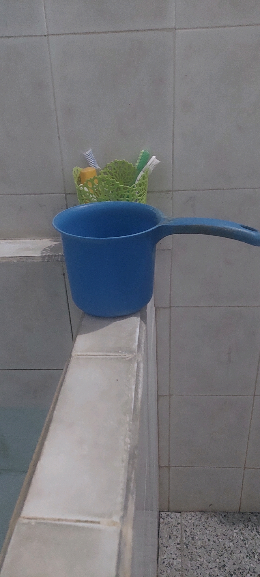 The image shows a blue plastic dipper resting on the edge of a bathtub or sink in a tiled bathroom. Behind the dipper, a green plastic holder contains several toothbrushes. The environment appears to be a residential bathroom in Lumajang, Indonesia. The overall lighting suggests an indoor setting, possibly during the daytime.