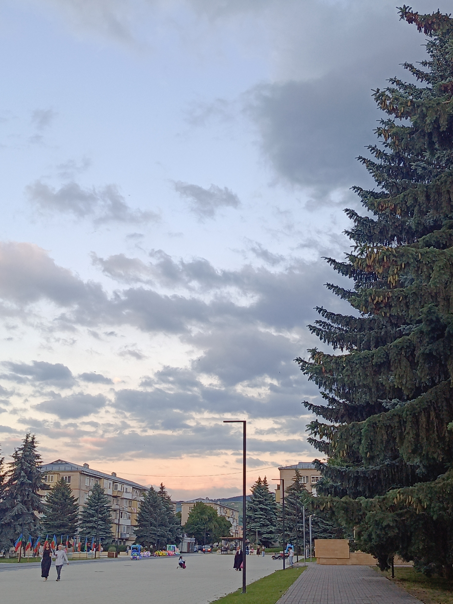 The image depicts an outdoor scene in Ust-Dzheguta, Russia, featuring a pedestrian walkway and a public square. A large coniferous tree with cones dominates the right side of the frame, its branches reaching towards a sky filled with scattered clouds. In the background, several multi-story apartment buildings line the square, interspersed with more coniferous trees. A street lamp stands on the left side of the walkway. Several individuals are visible in the square: two women are walking together on the left side of the walkway, and a child is riding a small vehicle in the distance. There are also some colorful amusement rides present in the square. The overall lighting suggests it is either early morning or late afternoon, with a soft, diffused light. The pavement of the walkway is made of rectangular bricks.