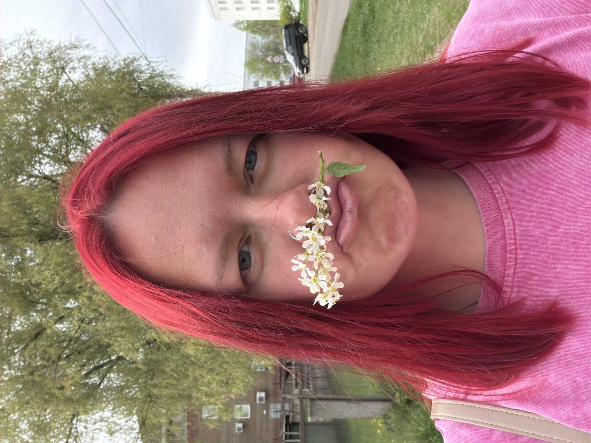 A woman with red hair wears a pink shirt and presses a flowering branch to her face. The background includes trees, a building with windows, and a street. The location is in the city of Chaikovsky, Russia.