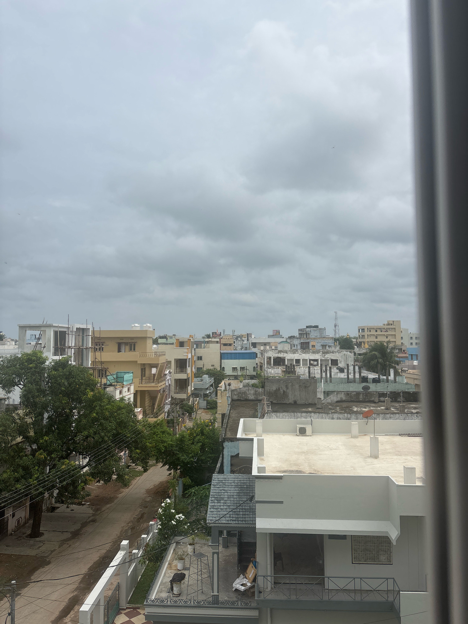 The image depicts a view of a city with overcast skies, likely in Srikakulam, India. The scene is dominated by multi-story buildings of various architectural styles and colors, some appearing to be residential homes and others possibly commercial structures. Trees, including a large one with dense foliage on the left, are interspersed among the buildings. A road runs through the lower left portion of the image, with some scattered debris and a parked vehicle visible. Several power lines are strung between poles. The rooftops of many buildings are visible, some featuring water tanks, satellite dishes, and air conditioning units. The overall impression is of a densely populated urban environment.