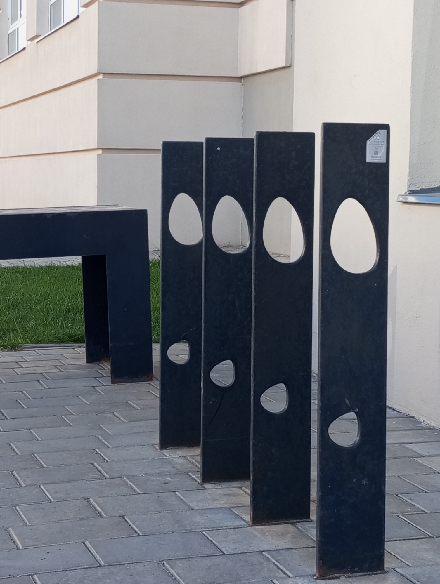 The image depicts a row of four black, rectangular metal pillars with oval and teardrop-shaped cutouts. These pillars are likely a public art installation or a modern bicycle rack. A black metal bench sits to the left of the pillars, partially obscured by the foremost pillar. The background features a light-colored building with windows and a brick-paved walkway. The presence of a QR code sticker on the last pillar indicates this is located in a public area. Based on the prompt, the scene is in the city of Pervomaysky, Russia. There are no people visible in the image. The environment is clean and well-maintained.