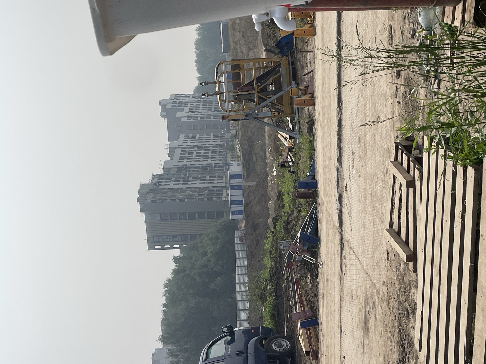 The image depicts a construction site in Koltsovo, Russia, characterized by a hazy sky and unfinished buildings in the background. In the foreground, a blue truck is parked next to a pile of construction materials, including wooden planks and metal components. A yellow metal structure, possibly part of a lifting device, stands prominently in the middle ground. Several portable toilet units are visible in the distance. The ground is a mixture of dirt and paved surfaces, with vegetation growing around the edges. The overall scene suggests ongoing construction activity in a developing area.