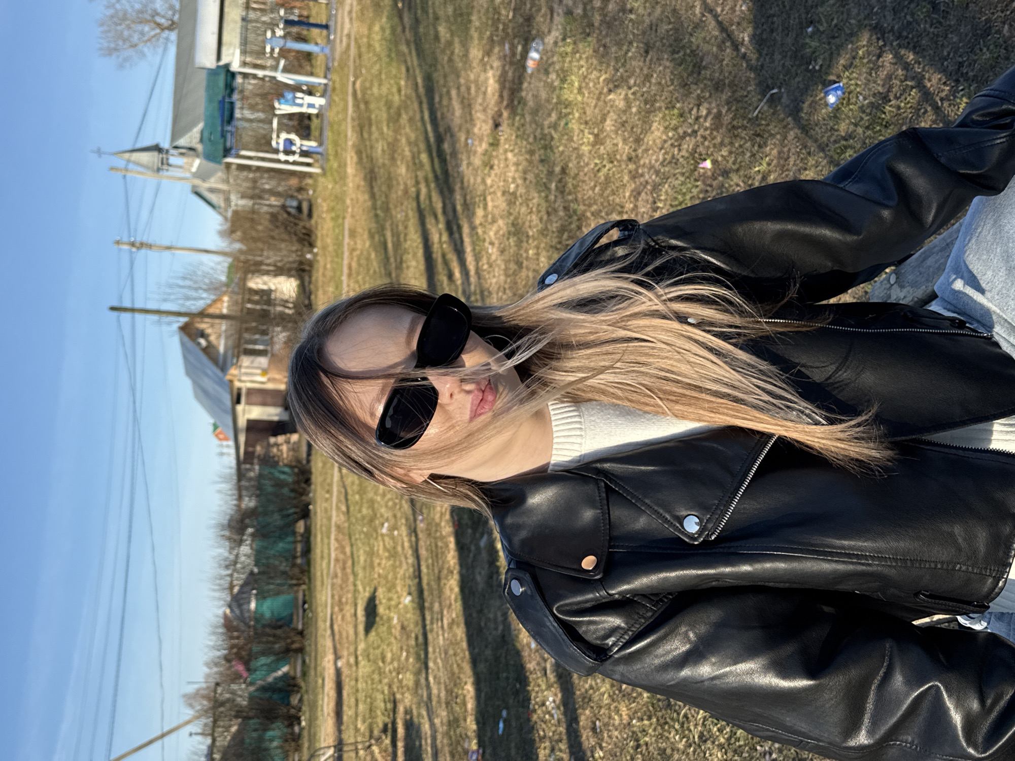 The image shows a young woman posing outdoors on a sunny day in what appears to be a rural or village setting, likely in Фоки, Russia. She is positioned in the foreground, captured in a profile-like pose, looking towards the right side of the frame. She wears dark sunglasses, a black leather jacket over a white ribbed sweater, and light-colored jeans. Her long, light brown hair is swept across her shoulders and face, suggesting movement or wind. Her lips are slightly parted.
The background features a clear blue sky and a sparse, dry grassy field littered with various pieces of debris, including plastic bottles and wrappers. In the middle distance, several wooden utility poles with wires run across the landscape. On the left side of the frame, buildings are visible, including one with a distinct steeple and cross, resembling a church. Further in the background, blue outdoor exercise equipment can be seen. Bare trees and bushes are scattered throughout the scene, suggesting either early spring or late autumn. The strong sunlight casts shadows, indicating mid-morning or late afternoon. No discernible text is visible on any objects or structures.