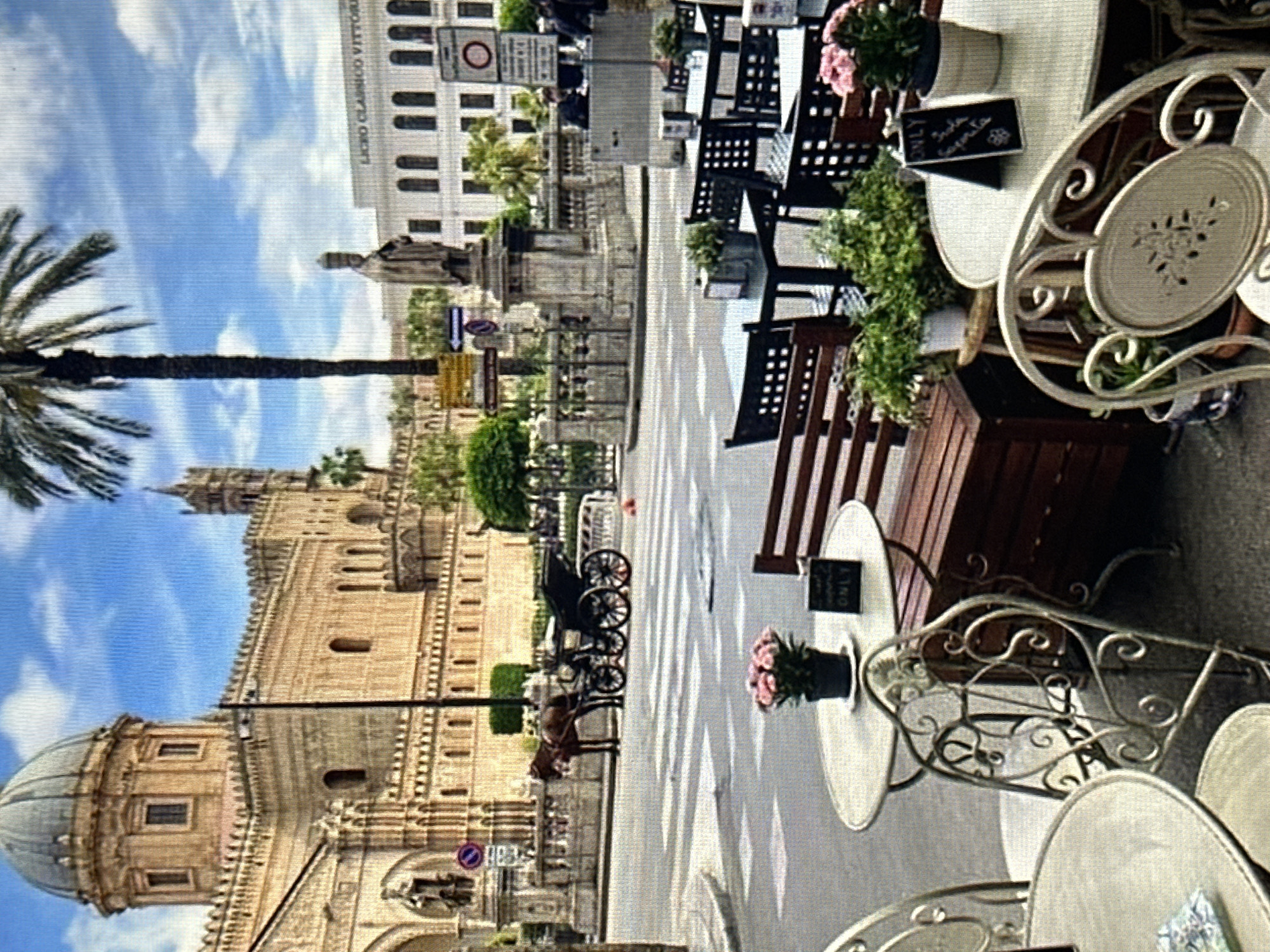 In an outdoor urban setting with a focus on architecture and street life, a horse-drawn carriage is stopped at a street crossing in front of a grand building with a dome and intricate stonework. The building appears to be a cathedral or a similar historical structure. In the foreground, several tables and chairs are set up, suggesting a cafe or restaurant. Some of these tables have potted plants and small signs. A tall palm tree stands prominently in the background. The sky is blue with scattered clouds. The overall atmosphere is that of a European city, likely in a warm climate, with a blend of historical and modern elements.