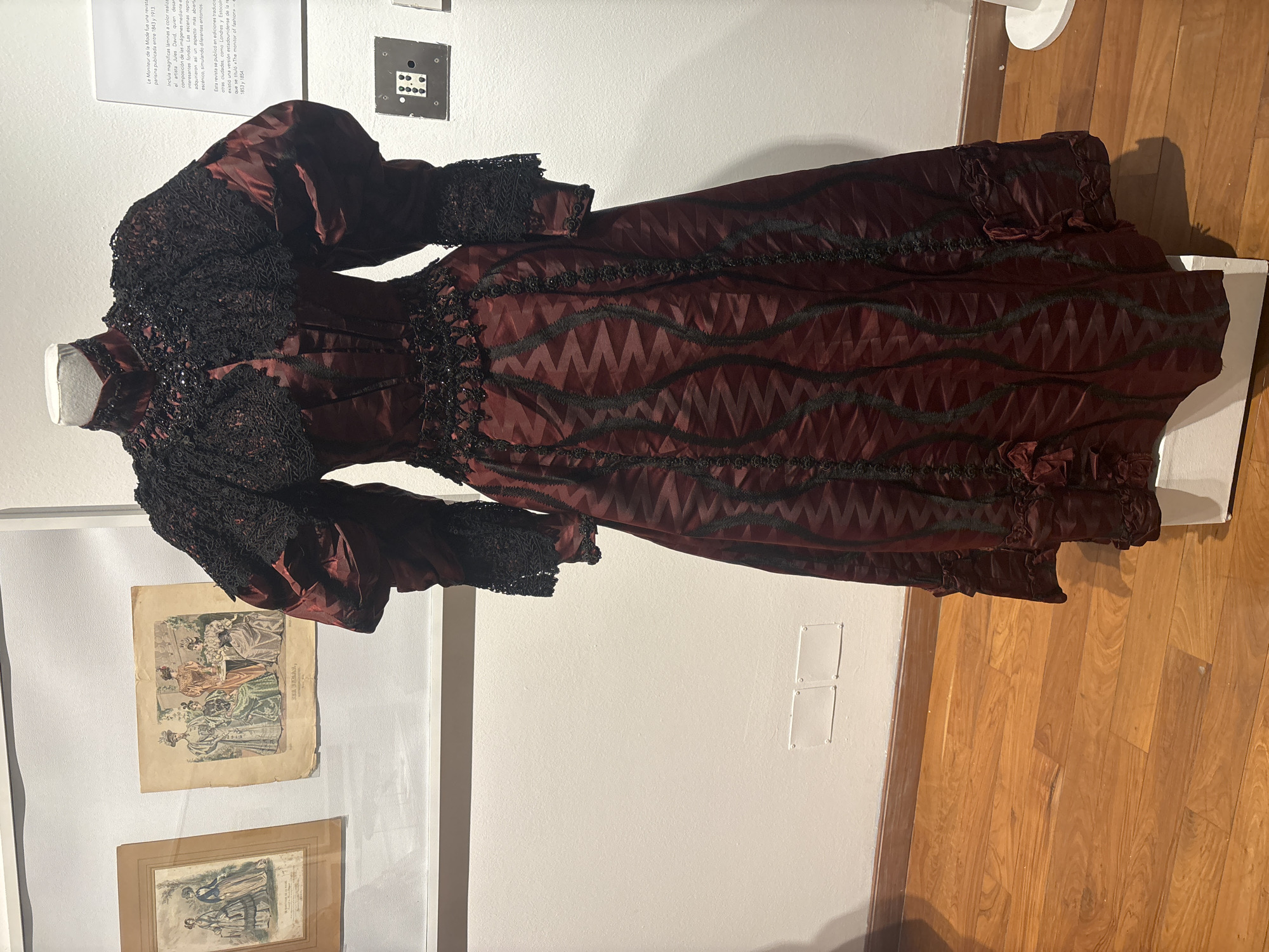 A museum exhibit features a maroon and black Victorian dress displayed on a mannequin. The dress has lace detailing around the neck and sleeves, and a patterned skirt. Behind the dress are framed illustrations and text panels, likely describing the dress and its historical context. The exhibit is located in Buenos Aires, Argentina, as evidenced by the Spanish text on the descriptive panel. The floor is wooden.