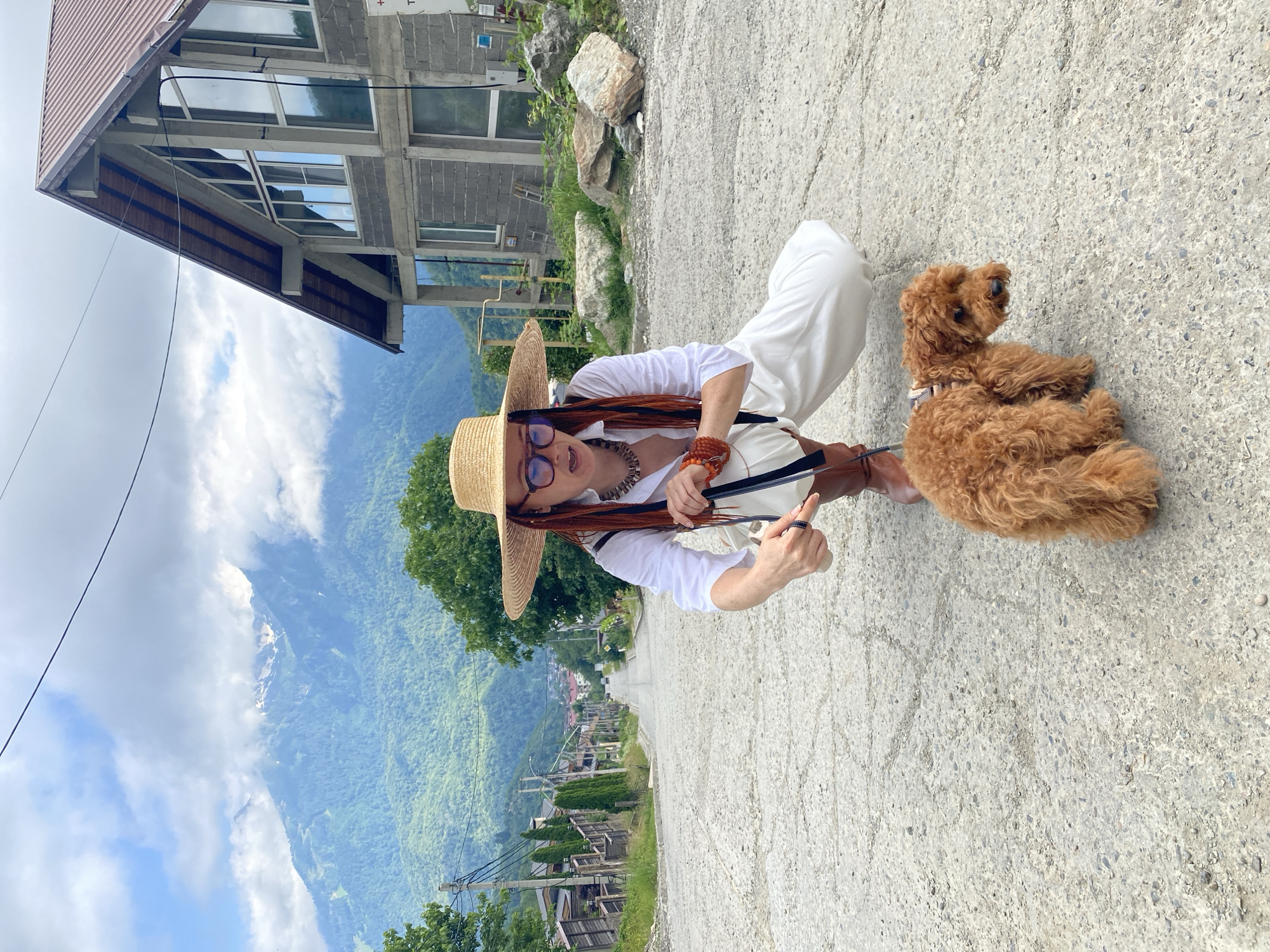 A woman with long, red hair, wearing a wide-brimmed straw hat, glasses, a white button-down shirt, and white pants, is crouching on a steep, unpaved road in Krasnaya Polyana, Russia. She is holding a leash attached to a small, reddish-brown poodle. The dog is standing on the road in front of her. In the background, a mountain range covered in green trees and some clouds is visible. A modern, unfinished house with large windows and a metal roof is situated to the right of the woman. The road slopes downwards, with houses and trees lining the sides. The sky is partly cloudy.