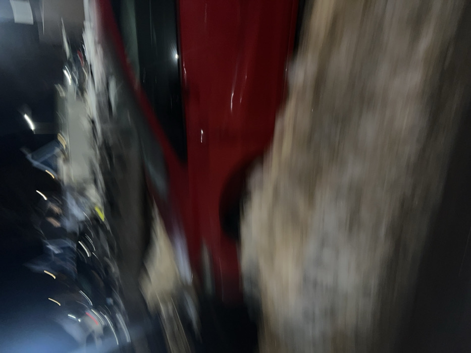 The image displays a blurry, low-light nighttime scene in New Haven, United States of America, characterized by significant motion blur. A red sedan, occupying the mid-ground, appears positioned against or partially within a substantial accumulation of dirty, partially melted snow or slush. The immediate foreground consists of a dark surface, likely asphalt, partially covered by the snow accumulation. In the highly blurred background, multiple vehicles are discernable, suggesting a commercial automotive environment such as a dealership or a large parking lot. Elongated streaks of artificial light sources, consistent with streetlights or building illumination, are visible across the distant background. No individuals are clearly identifiable within the frame. The scene depicts winter conditions. The precise street address or specific landmark within New Haven is not discernible from the image, but the setting is consistent with a commercial or industrial zone.