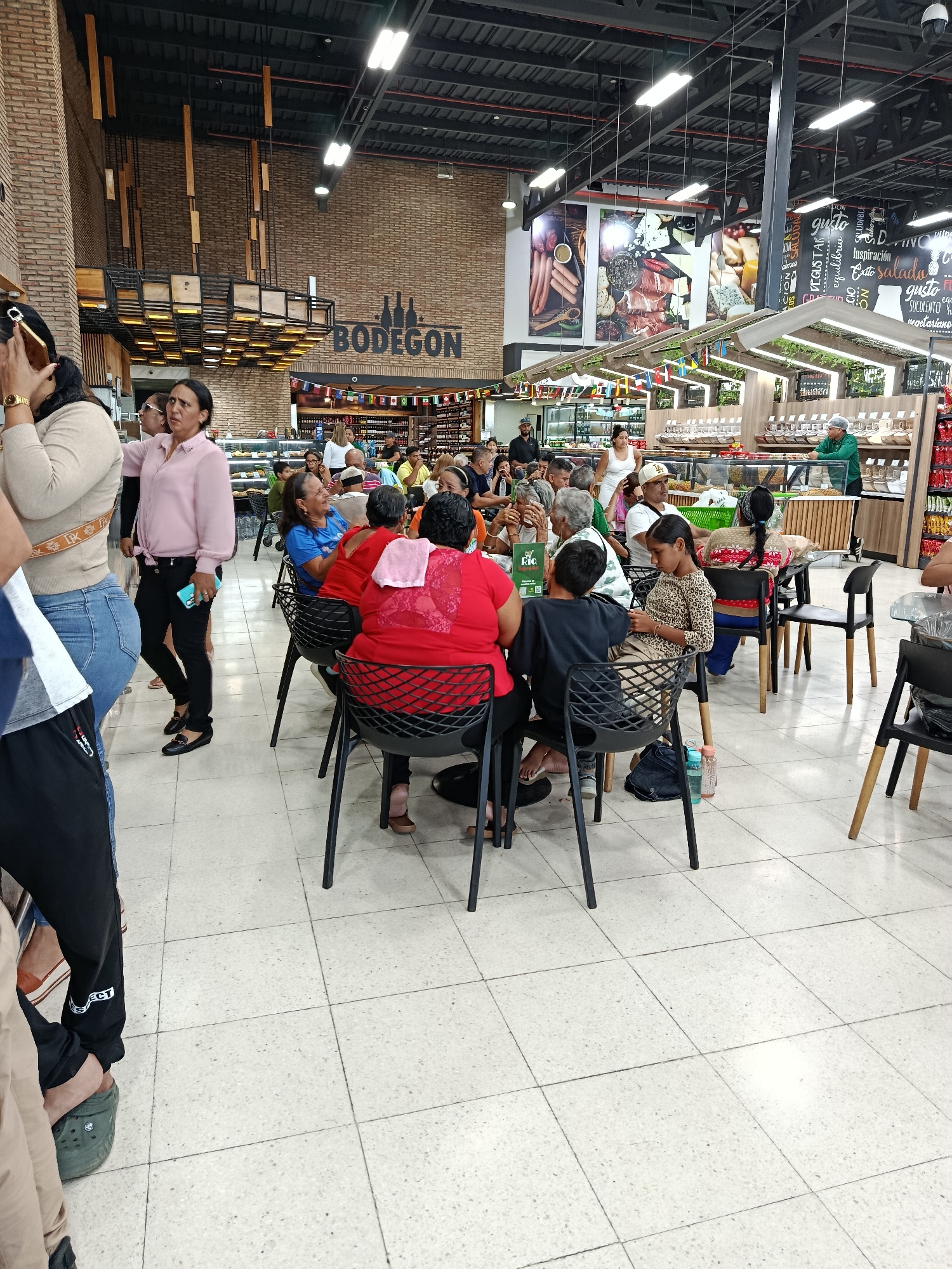 The image depicts the interior of a busy market or grocery store, possibly a supermarket, in Juan Griego, Venezuela. In the foreground, several people are seated at tables, seemingly in a dining area within the establishment. One person, wearing a red shirt, is seen from the back, with a pink cloth draped over their shoulder. Another person, a young girl in a leopard print top, is seated at a table looking at her phone. There are chairs and tables arranged in a dining space. In the background, there are shelves stocked with various goods, and display cases. The overall environment appears to be a commercial space with bright lighting. The word "BODEGON" is visible on a brick wall, which is a common term for a warehouse or a large store in Spanish-speaking countries. Several people are standing and walking around, suggesting a lively atmosphere. The presence of flags hanging above some of the displays indicates a potential international aspect or promotion.