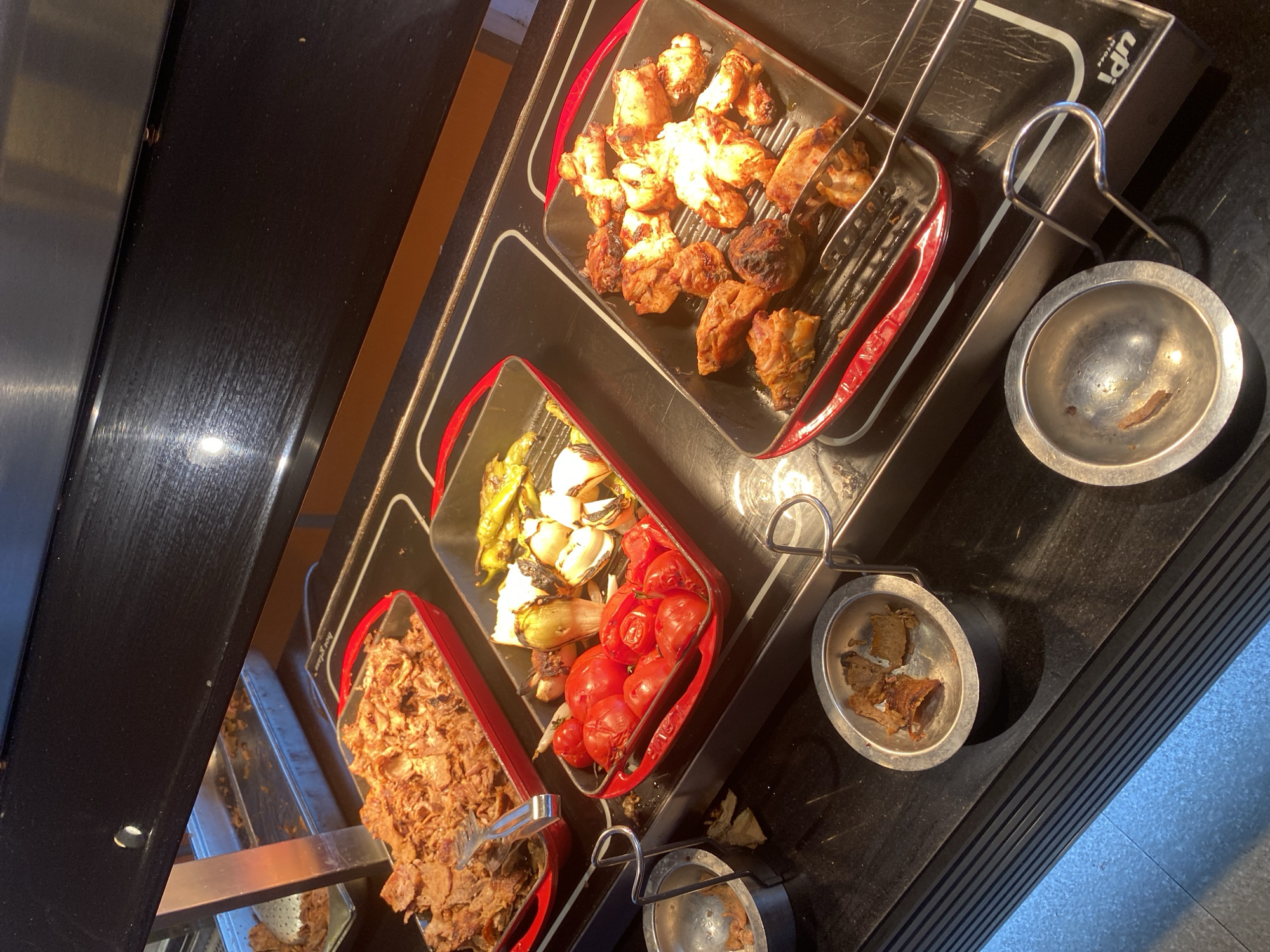 The image shows a food buffet display, likely in a restaurant or hotel. There are three rectangular metal pans with red edges, each containing different food items. The leftmost pan holds a generous portion of shredded meat, possibly döner kebab or shawarma, with metal tongs resting on top. The middle pan contains roasted vegetables, including what appear to be tomatoes, onions, and bell peppers. The rightmost pan displays pieces of grilled chicken. In front of the pans are two small silver bowls, one containing a few pieces of what looks like bread or pastry, and the other appearing empty. The buffet is illuminated by overhead lights, and the surfaces are dark and reflective. The overall scene is one of a self-service food offering. The text "upi" is visible on the right side of the image, possibly indicating the brand of the heating element. The location is identified as Aksu, Turkey.