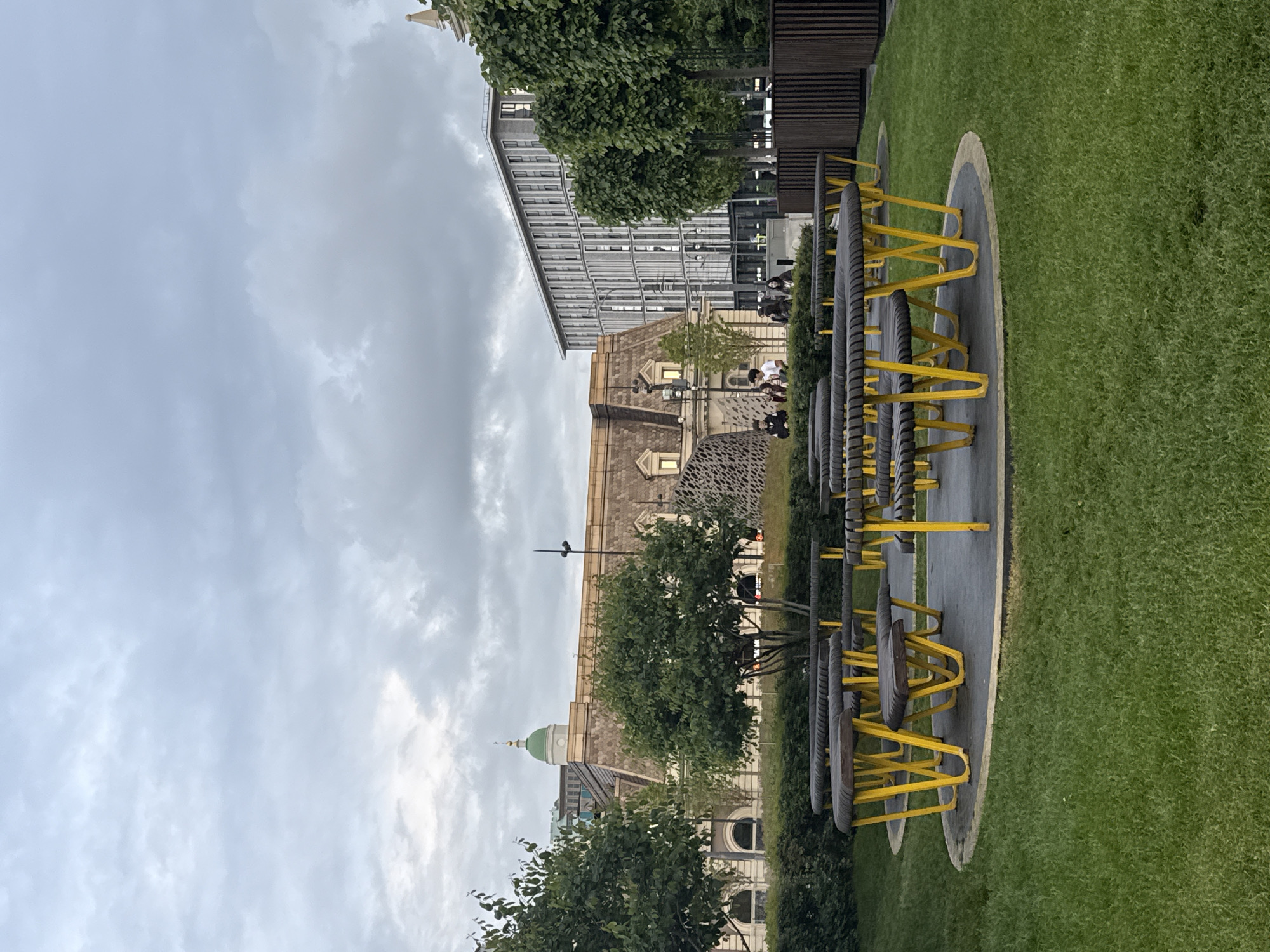 The image depicts a playground or park setting in an urban environment. There are rows of exercise equipment with yellow bars and dark tires or rollers along the ground on a gray surface surrounded by a border and a grass surface. In the background are buildings including one with a green dome, another of stone construction, and a more modern building with a glass facade. A few people are visible in the background, some sitting near the stone building. The sky is cloudy. Based on architectural styles and information, the location may be in Moscow, Russia.