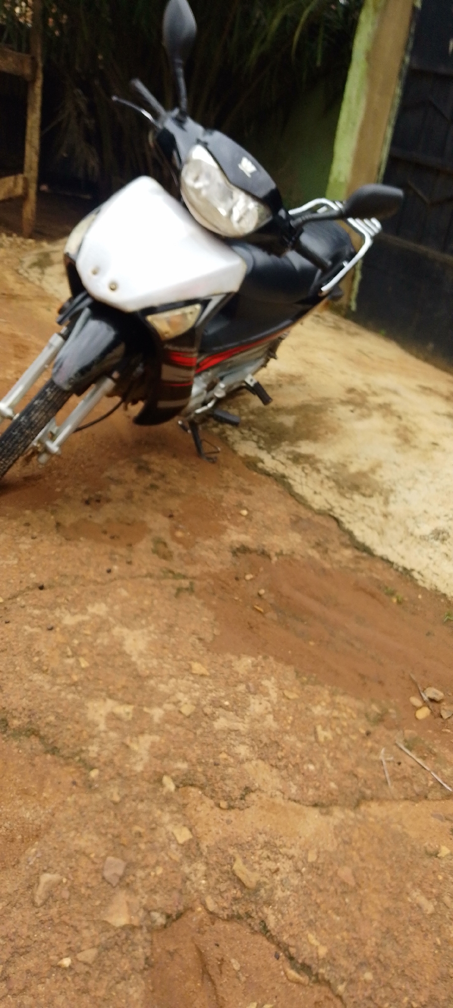 A black and white motorcycle is parked on a dirt road in Baissa, Nigeria. The motorcycle is a street bike model with a white front fairing, black body panels, and red accents. The front tire is visible, and the handlebars are turned to the left. The background consists of lush green foliage and a wooden structure. The ground is covered in reddish-brown dirt, with some patches of sand and small stones. A light-colored post with a dark top is visible to the right of the motorcycle. The scene appears to be outdoors, possibly in a rural or semi-rural area.
