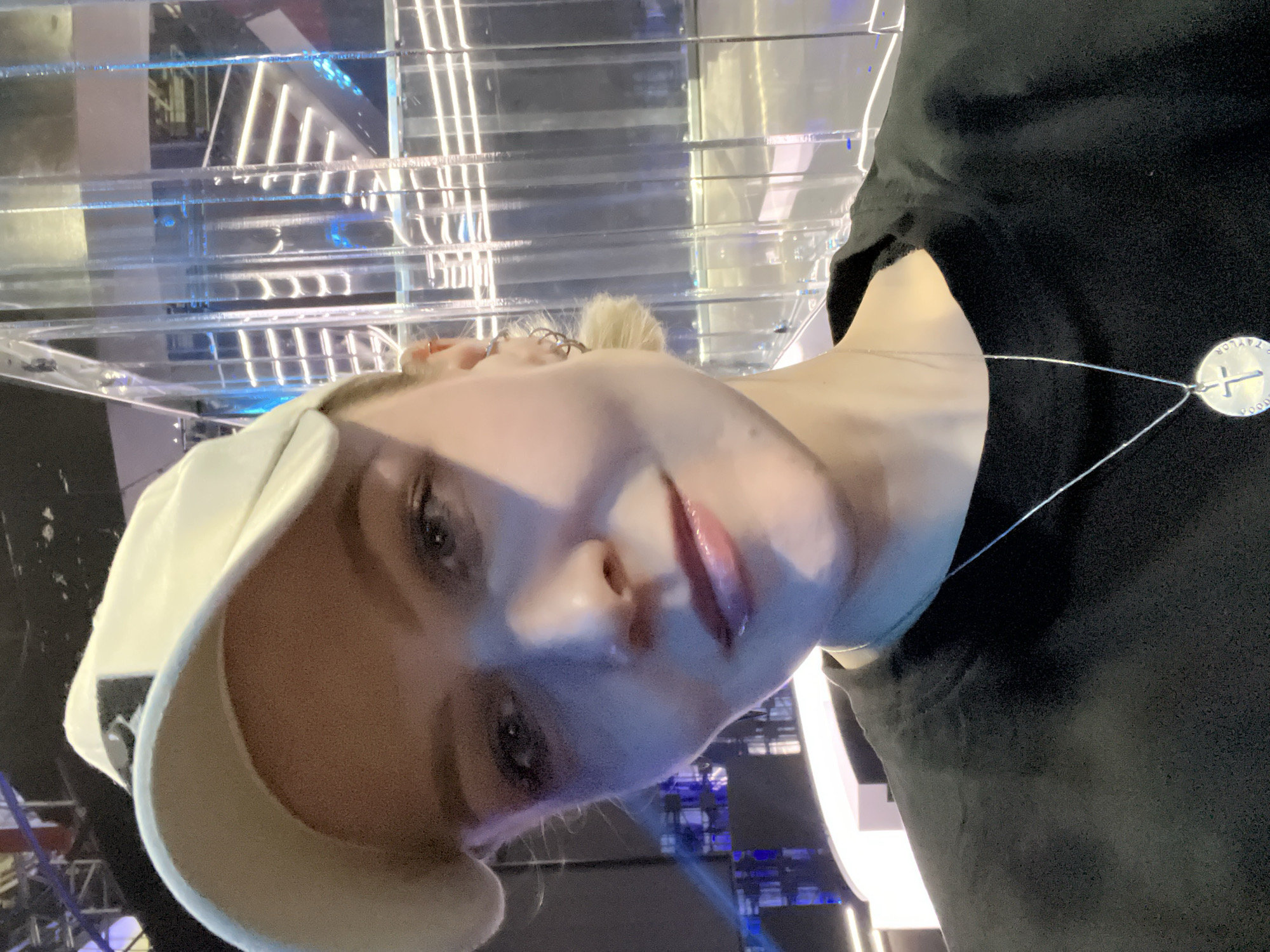 A close-up portrait features a person wearing a white baseball cap and a dark, possibly black, long-sleeved shirt. The individual has light-colored hair, pulled back into a bun or ponytail visible in the background crops. Their face is illuminated by artificial light, highlighting their eyes and glossy lips, suggesting a neutral to slightly serene expression as they look directly into the camera.
A silver circular pendant hangs from a thin chain around their neck, visible over the dark shirt. The pendant features an engraved cross in the center, and text encircling it. The legible text includes "TAYLOR" on the bottom left side, "POODE" on the right side, and a word that appears to be "NOVAYA" or "NOTAYA" on the top left segment.
The background consists of a modern, indoor architectural setting. Numerous clear or translucent panels, likely glass or acrylic, are stacked or arranged in parallel, creating horizontal lines and a sense of depth. Behind these panels, linear light fixtures, possibly LED strips, cast a cool, blue-white light, giving the scene a structured, almost futuristic aesthetic. The lighting suggests an artificial, possibly nighttime, indoor environment, such as an exhibition hall, a modern museum, or a commercial space. No specific activity beyond posing for the photograph is evident. The image is taken at an oblique angle, adding a dynamic feel to the composition.
