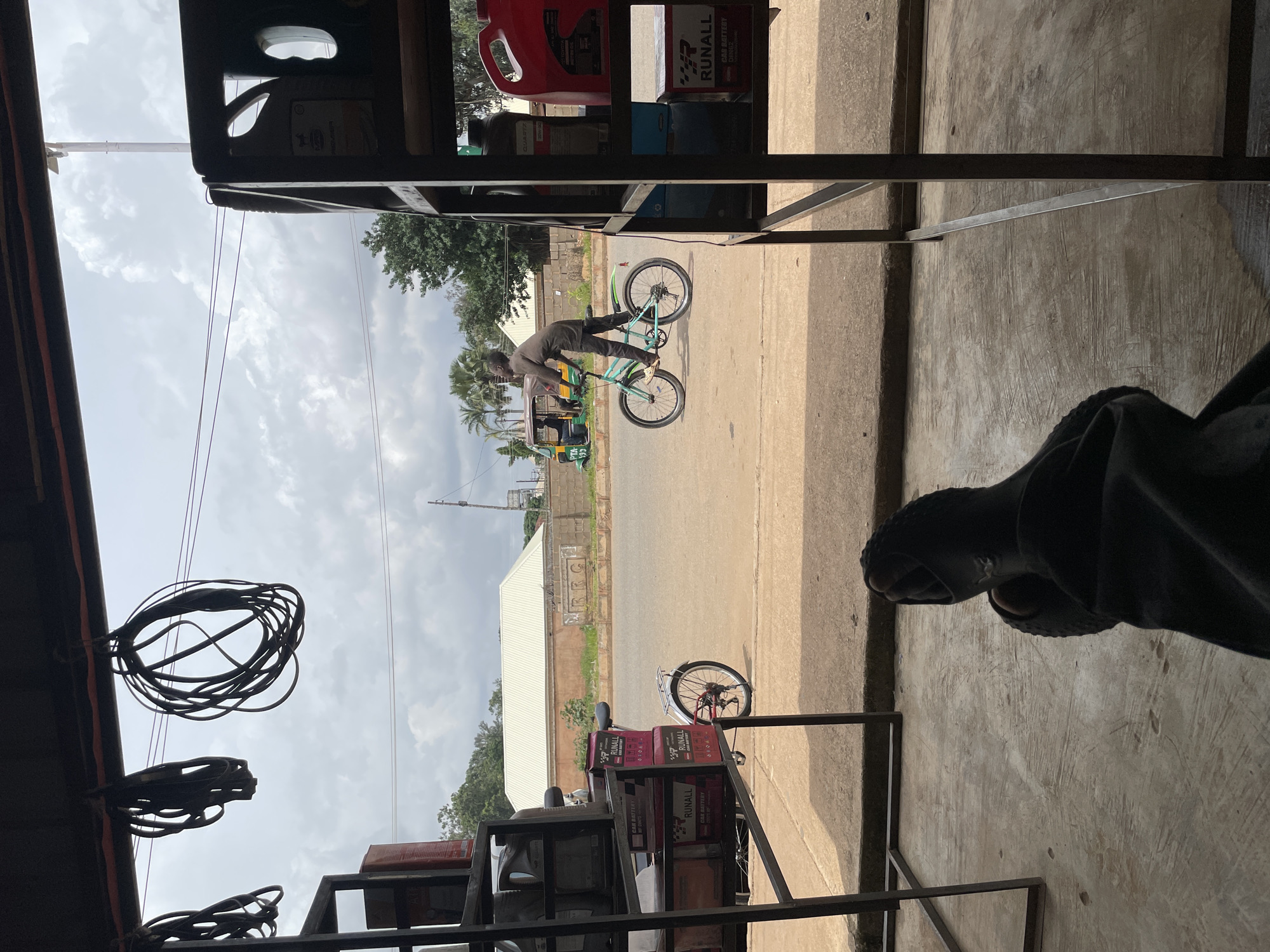 The image captures a street scene in Bukuru, Nigeria, viewed from inside a shop or open-fronted establishment. In the foreground, a person's legs and feet, clad in dark pants and open-toed sandals, are visible, suggesting they are sitting or reclining. The floor is concrete.
In the mid-ground, a young person is riding a turquoise bicycle on a paved road. The rider wears dark clothing. To the left of the road, a red and white bicycle is parked. Behind the bicycles, a small, three-wheeled vehicle, commonly known as an auto-rickshaw or Keke Napep, is visible with two occupants. Across the road, there are buildings with light-colored roofs and some trees. The sky is overcast with scattered clouds.
Within the shop's interior, metal shelving units are present. One shelf displays several red and black boxes labeled "RUNALL CAR BATTERY." Other shelves hold various containers, some appearing to be oil or fluid canisters, including a large red one. Hanging from the ceiling are several coiled black rubber belts or hoses. The overall impression is of a commercial area, possibly an auto parts or service shop, on a cloudy day in Bukuru.
