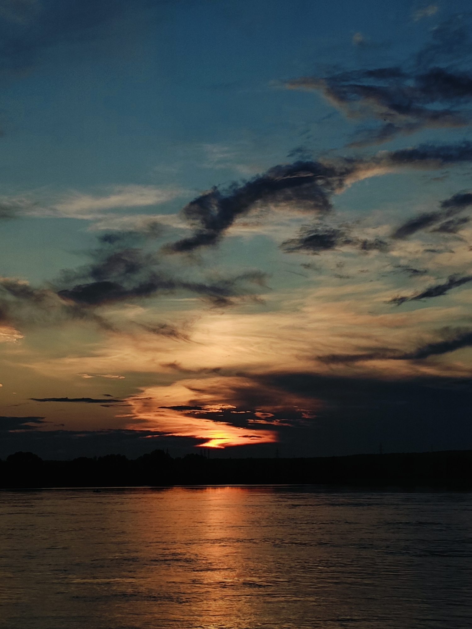 The image depicts a serene sunset over a wide body of water, likely the Kama River in Chaikovsky, Russia. The sky is a canvas of twilight colors, transitioning from deep blue at the top to hues of orange, pink, and yellow near the horizon. Wispy clouds, illuminated by the setting sun, add texture and depth to the sky.
The sun itself is a fiery orange orb, partially obscured by dark clouds, casting a warm, shimmering reflection on the water's surface. The water is calm, with gentle ripples distorting the reflection of the sky and the silhouette of the distant shoreline.
Along the horizon, a dark, silhouetted treeline marks the edge of the land. The dark band of trees suggests a forested or rural area. In the distance, faint outlines of what appear to be man-made structures, possibly industrial or power line towers, can be discerned against the fading light.
In the foreground, the water occupies a significant portion of the image. The surface is predominantly dark, reflecting the muted tones of the sky. There is a small, dark object on the water, barely visible, which could be a boat or a raft, hinting at human presence. However, there are no clearly discernible individuals or specific actions taking place.
The overall atmosphere is one of tranquility and natural beauty, characteristic of a peaceful evening by the river. The image captures a moment of transition between day and night, with the vibrant colors of the sunset creating a dramatic and visually appealing scene.