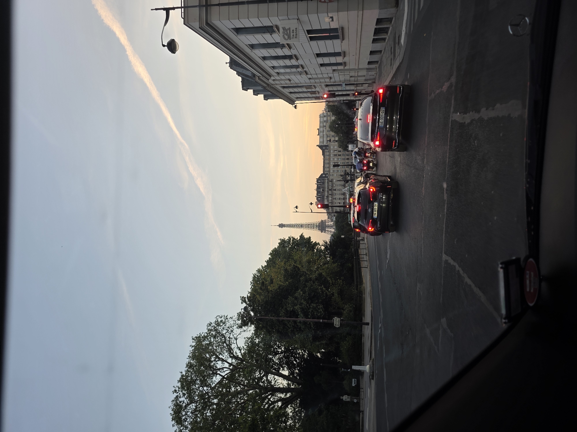 The image captures a street view in Paris, France, at dusk. The iconic Eiffel Tower is visible in the distance, silhouetted against a sky with a faint contrail. In the foreground, traffic is stopped at a red light. Several cars, including two black sedans, are on the road. A building with a French bank logo ("Crédit Agricole Ile de France") is on the right side of the frame. Large trees flank the left side of the image, and a street lamp is present. The scene appears to be a typical urban intersection with moderate traffic flow during evening hours.