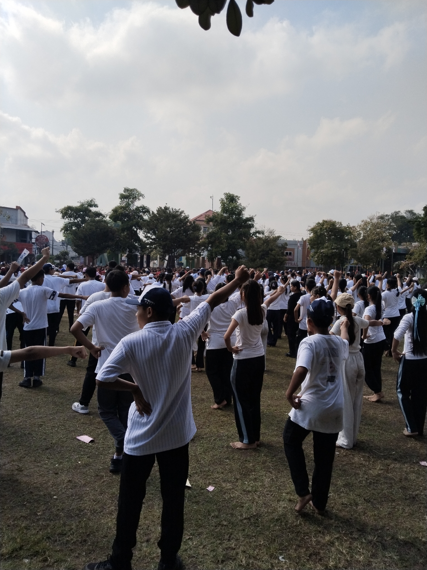 In the city of Kecamatan Pati, Indonesia, a large group of people, primarily young individuals wearing white t-shirts and dark pants, are engaged in an outdoor group exercise or dance session on a grassy field. The sky above is overcast with scattered clouds. Several trees are visible in the background, along with buildings that appear to be residential or commercial structures. Some individuals are barefoot, while others wear athletic shoes. The participants are arranged in rows and appear to be following coordinated movements, with many raising their arms. The overall scene suggests a community event or a school activity taking place in a public space.