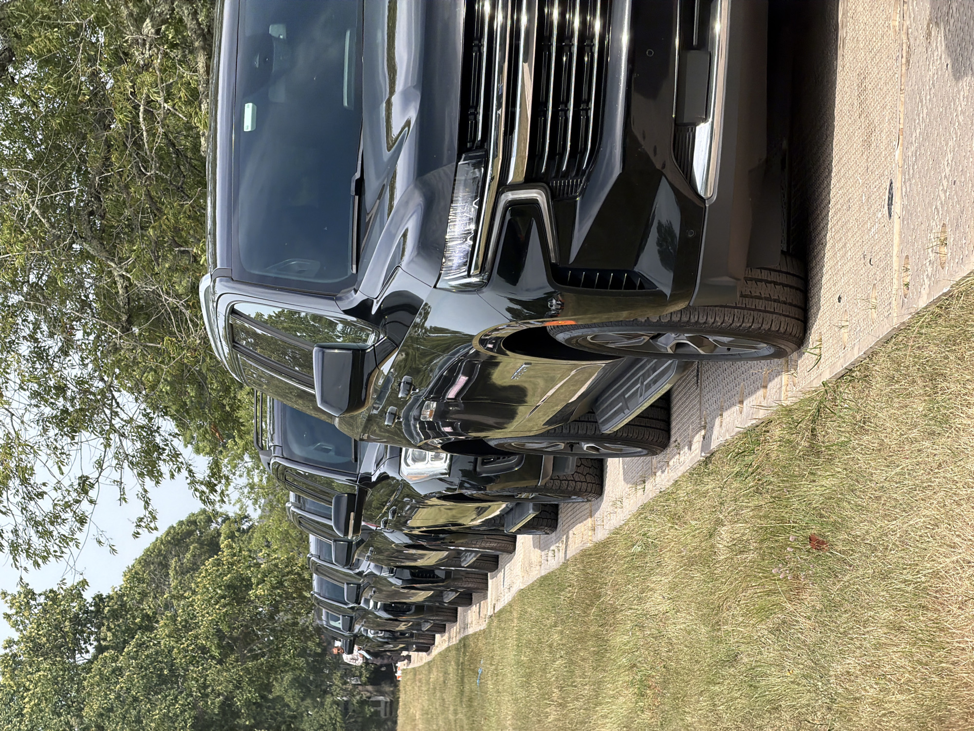 A line of black SUVs is parked on a textured surface next to a grassy area under a canopy of trees. The perspective is low, focusing on the front of the closest SUV, which has chrome accents on its grille and bumper. A man wearing a light-colored patterned shirt and dark pants stands in the background, looking down at something in his hands, positioned to the left of the parked vehicles. The environment appears to be outdoors, possibly a park or a roadside, with dry grass and lush green trees. The sky is visible through the branches, suggesting it is daytime. The scene is in the Town of Southampton, United States of America.