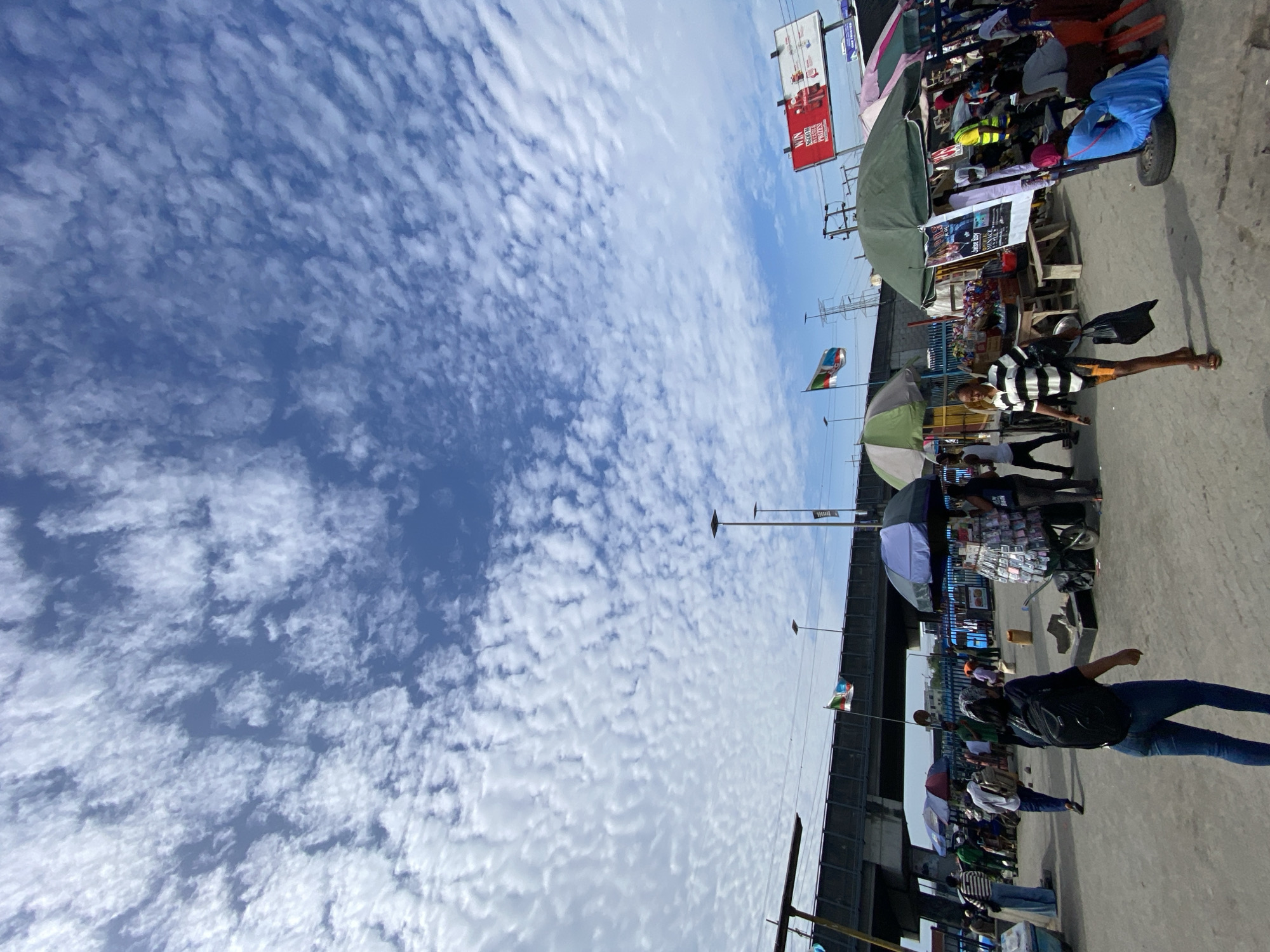 The image shows a street scene in Lekki, Nigeria, with a bustling market under an overpass. The sky above is filled with scattered cumulus clouds. People are walking and interacting with vendors selling goods from stalls and carts, many shaded by umbrellas. A billboard is visible, and the flags of Nigeria and another nation are flying near the overpass structure. The scene depicts typical urban street life in Lekki with commerce and movement.