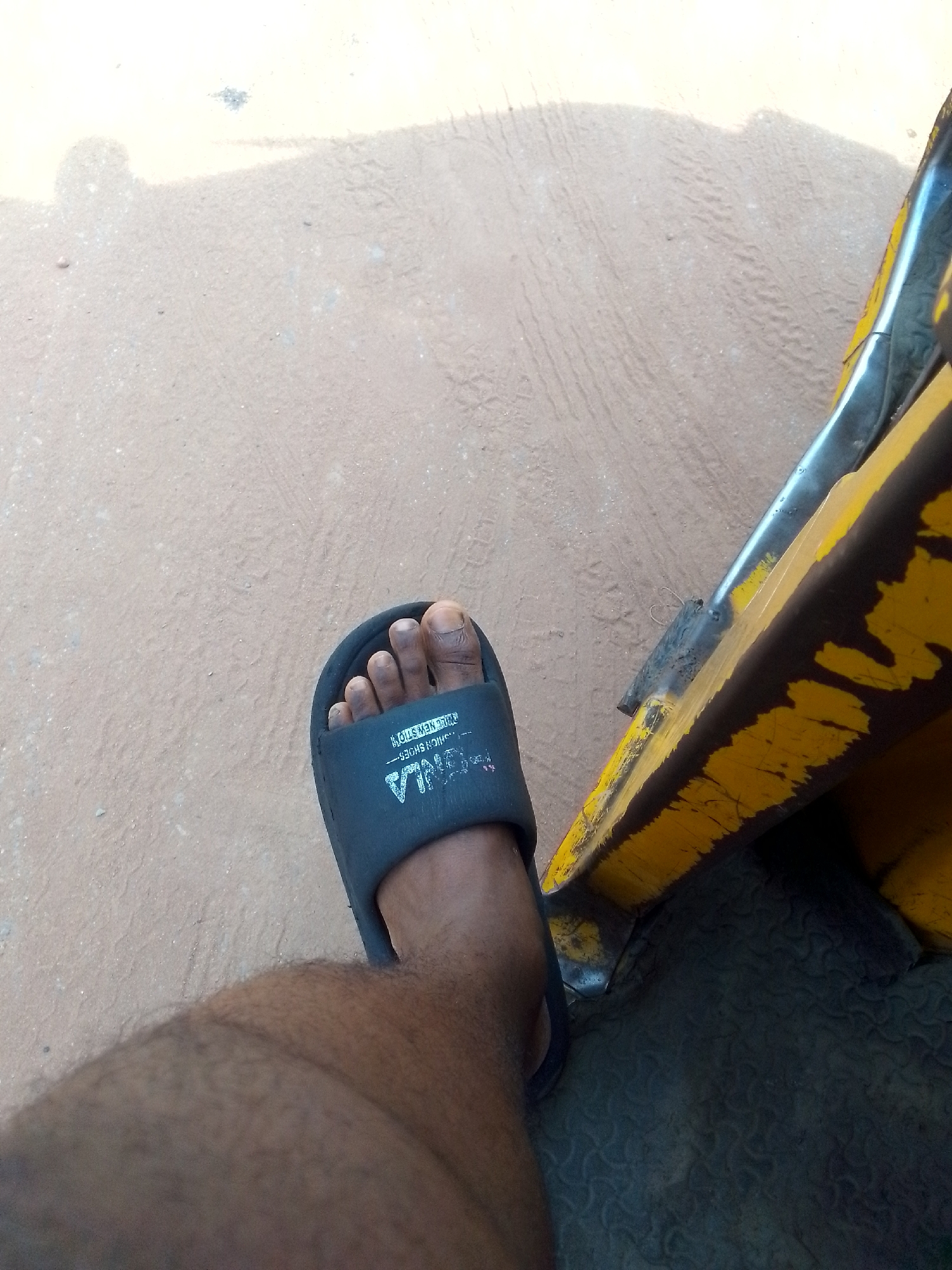 In Nkpor, Nigeria, an individual's foot, clad in a dark grey slide sandal, rests on a textured, light brown ground. The sandal displays white lettering, including the word "FASHION SHOES". To the right of the foot, a portion of a yellow vehicle is visible, characterized by worn paint and a dark band. The ground appears to be a dirt or gravel surface with tire tracks imprinted. The lighting suggests an outdoor setting, possibly during daylight hours.
