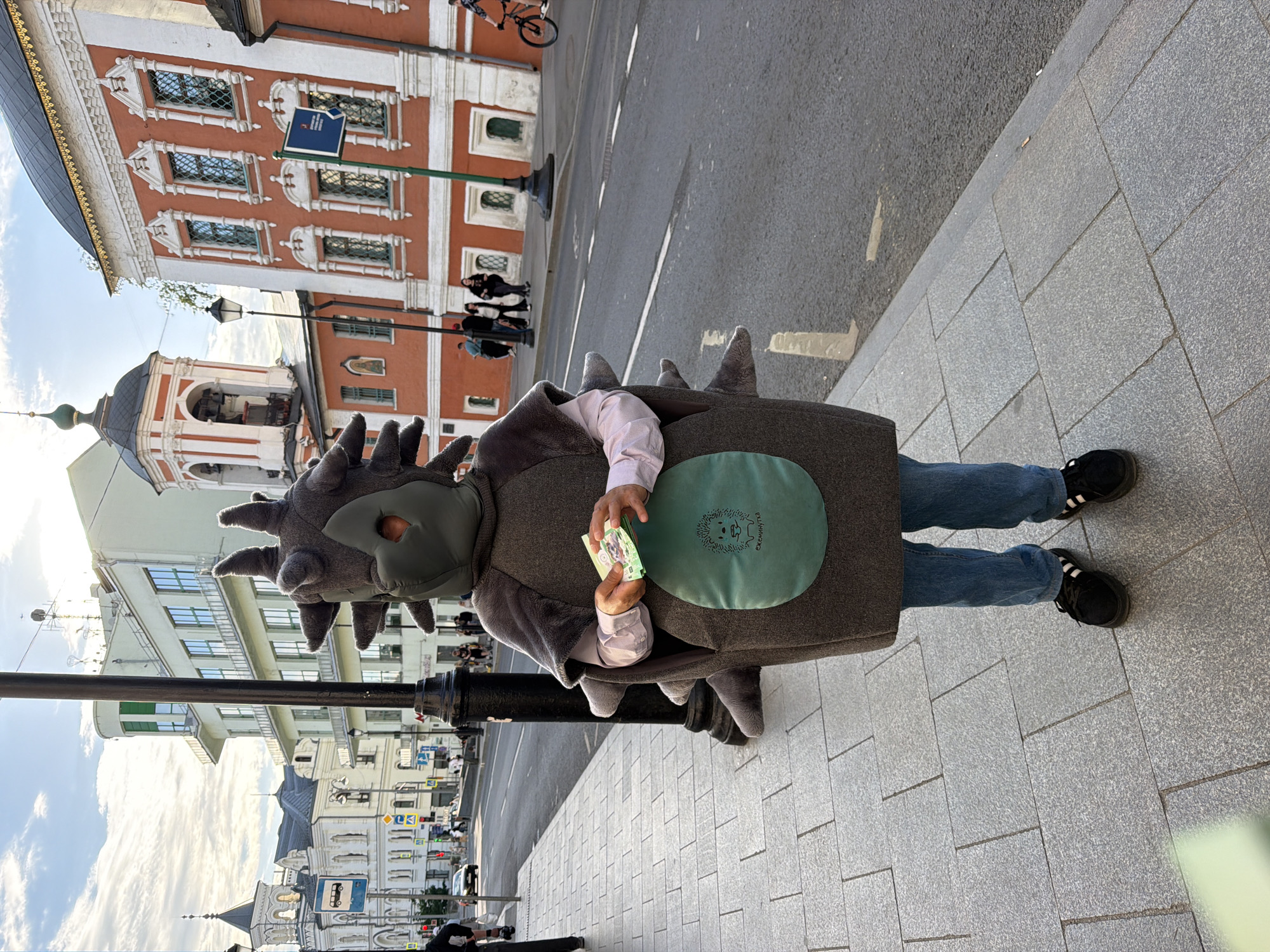 A person wearing a full-body hedgehog costume with spikes and a green circle on the chest is standing on a gray brick sidewalk. The person is wearing blue jeans and black sneakers. The person's hands are visible, holding a stack of paper money. The backdrop shows a street in Moscow, Russia, with buildings of various architectural styles, including some with orange facades and white trim, as well as a church with a bell tower. A black lamppost is in the foreground, and pedestrians are visible in the distance.