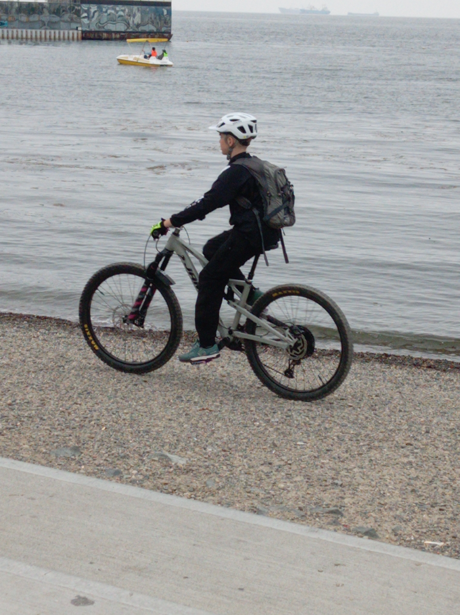 A person wearing a white helmet and a black jacket with a grey backpack is riding a grey bicycle on a gravel surface next to a body of water. The bicycle has black tires with yellow lettering and pink and black suspension forks. The person's left foot is on the pedal and the right foot is near the ground. In the background, there is a yellow boat with two people in it, a large structure on the left, and ships on the horizon. The scene appears to be near the waterfront in Vladivostok, Russia, possibly along the city's coastline.