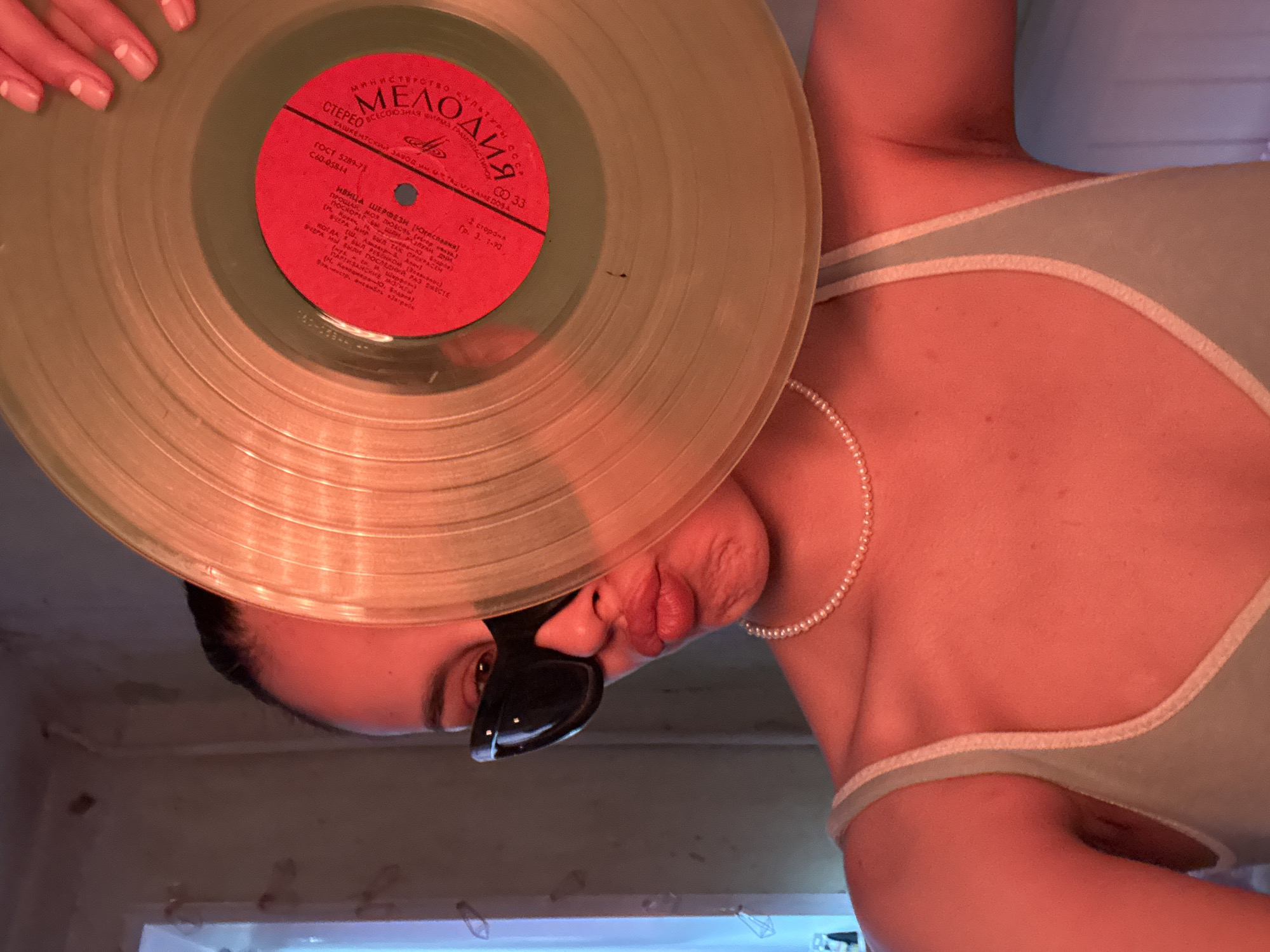 A person wearing a light blue tank top and a pearl necklace holds a translucent gold vinyl record in front of their face, obscuring it with the exception of their eyes, nose, and pursed lips. They are also wearing black cat-eye sunglasses. The background appears to be an indoor setting with a plain wall and a window with a blue light emanating from it. The lighting in the image is predominantly warm, casting a reddish-orange hue over the scene. There are no discernible landmarks or features that would indicate the location in Votkinsk, Russia.