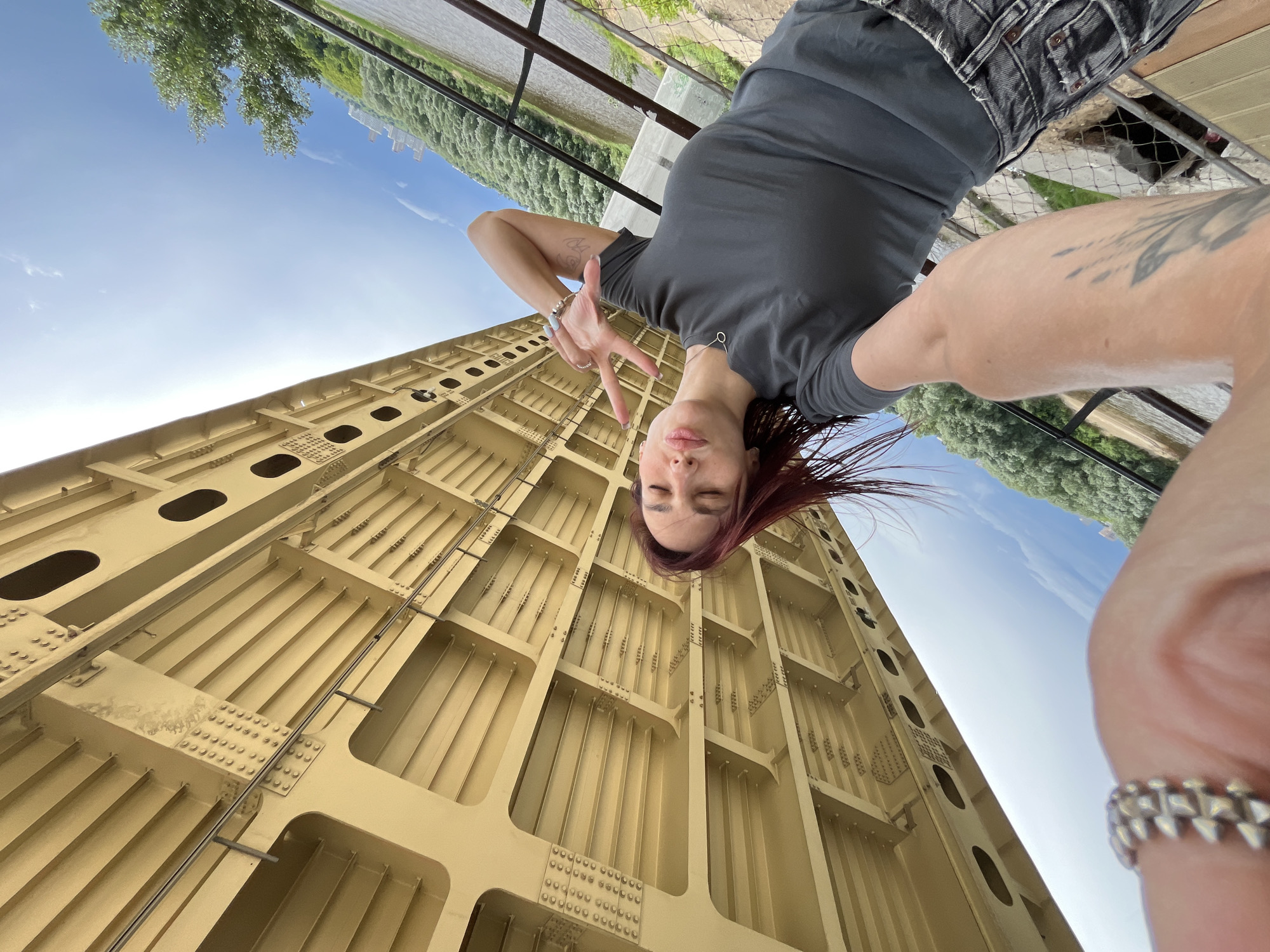 A woman with red hair takes a selfie from a low angle beneath a large, yellow, metal bridge structure. The bridge's underside is visible, showing its intricate beams and bolted joints. The woman is wearing a dark grey t-shirt and dark, possibly denim, shorts. She is making a peace sign with her right hand and has her left arm raised behind her head. Her eyes are closed, and her lips are slightly pursed. She is wearing rings on her fingers. In the background, to the right of the bridge, a body of water, possibly a river, is visible, bordered by lush green trees and hills. The sky is blue with scattered white clouds. The scene appears to be outdoors, likely in a park or recreational area adjacent to the river and under the bridge. It is difficult to definitively pinpoint the exact location within Nizhny Novgorod without further information, but the bridge and river setting suggest a location along the Volga River.