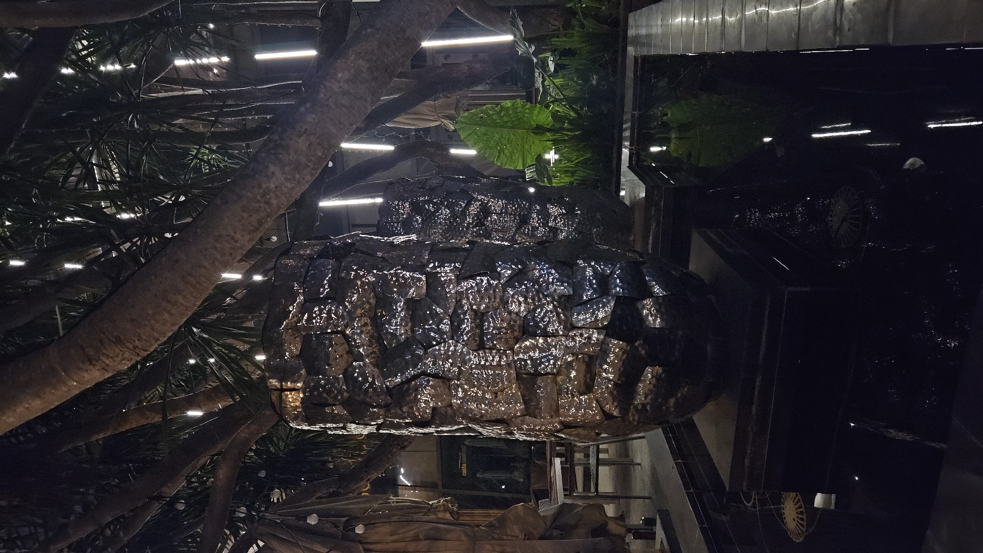 The image shows an indoor architectural feature in Nha Trang, Vietnam. A large, dark, metallic structure, possibly a decorative fountain or sculpture, is the central element. It appears to be layered, with a rough, textured surface. The scene is dimly lit, with artificial lights visible in the background, suggesting an indoor environment. Vegetation, including trees and large leaves, are present around the structure, indicating an attempt to integrate natural elements into the interior design. The background includes architectural details such as metal beams and what appears to be a building facade. A dark car, perhaps reflecting the lighting, is seen in the background. The overall scene suggests a modern, possibly upscale, indoor space in Nha Trang.
