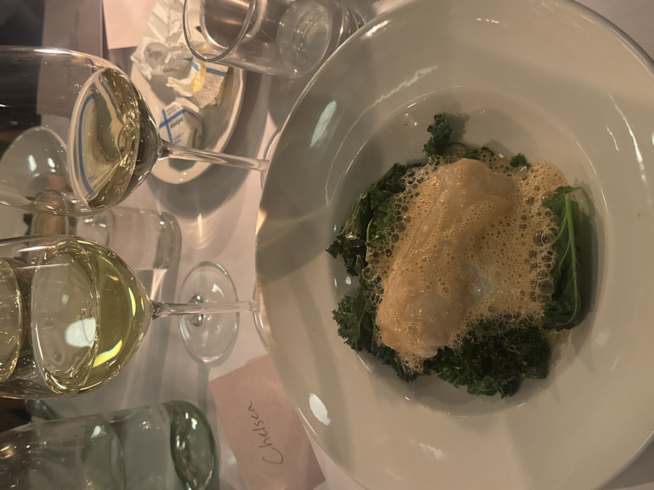 A plate of food presents fish atop green kale, enveloped in a brown, foamy sauce. Nearby, a stack of wine glasses, some containing pale yellow liquid, stands next to a small dish with butter packets. A light pink name card with "Chelsea" written on it is also visible on the table. The setting suggests a formal dining experience.