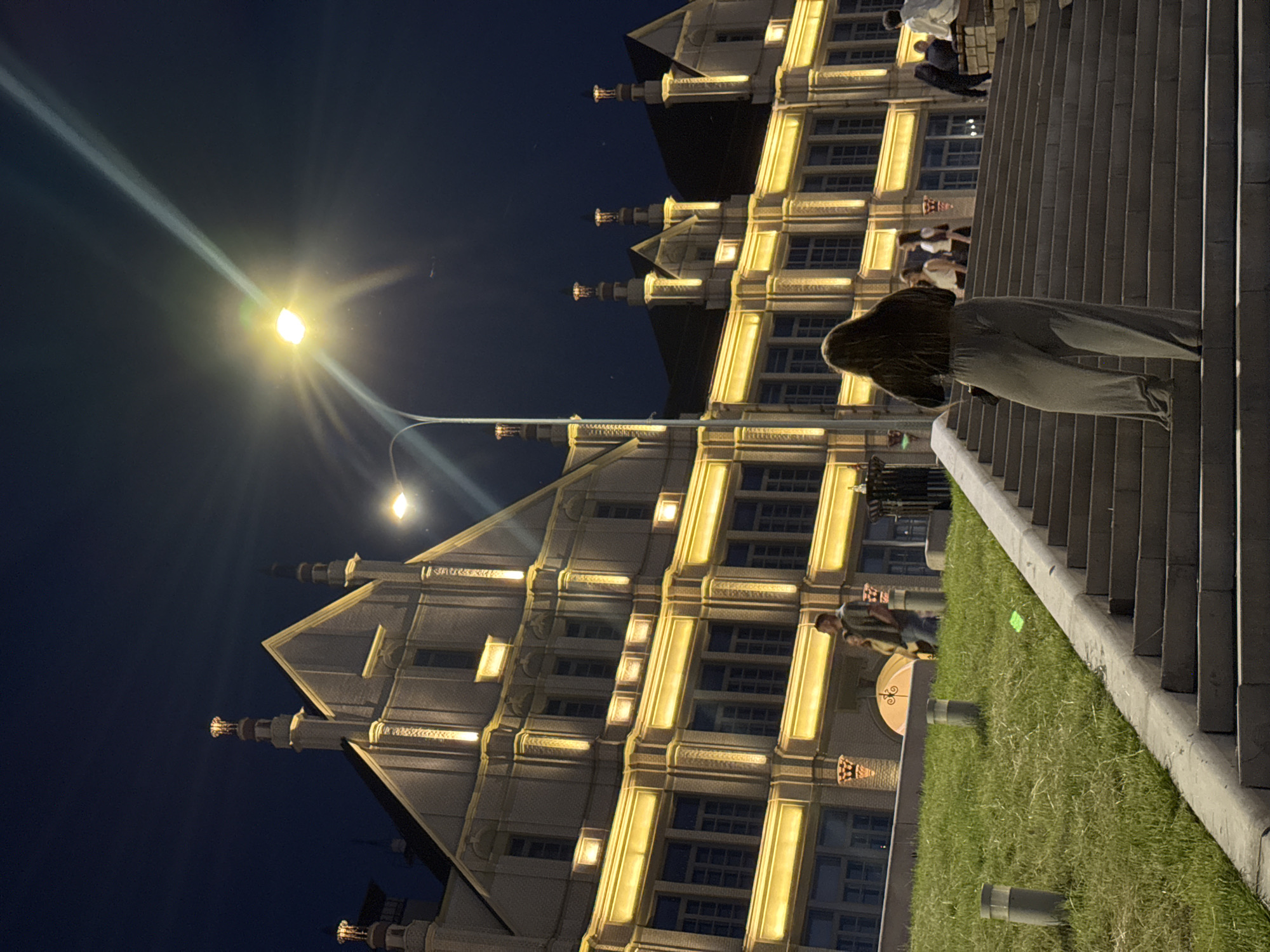 This image presents a nighttime scene in Nizhny Novgorod, Russia, focused on the illuminated Main Exhibition Building of the Nizhny Novgorod Fair complex. A tall, dark street lamp stands prominently in the mid-ground on the left, emitting a bright yellow light that generates a distinct diagonal lens flare extending upwards and to the right across the dark blue sky.
In the foreground, a wide, multi-tiered concrete staircase ascends from the bottom right towards the building. A person, viewed from behind, with long dark hair, wearing a dark top and loose-fitting light-colored trousers, is centrally positioned on these stairs, ascending. To the left of the staircase, a grassy slope is visible, dotted with three grey cylindrical bollard lights; a small, bright green object lies on the grass.
The primary subject is a large, multi-story building characterized by its grand architectural style. Its facade is a light, cream color, intricately detailed with prominent gables, decorative cornices, vertical pilasters, and multiple rows of windows. The building is extensively illuminated with warm yellow lighting that highlights its architectural elements, including the outlines of windows and decorative features. The roofline features several distinctive turrets or chimney-like structures, each capped with an illuminated finial.
Several other individuals are present within the scene. On a platform directly in front of the building, to the left, a man wearing a dark t-shirt and jeans walks beside a woman. Further up the stairs, behind the main ascending figure, two women are visible. To the far right, on a raised stone seating area, multiple additional figures are seated or standing, including a person in a white shirt. The overall environment is an urban setting, lit by artificial light sources under a dark night sky.