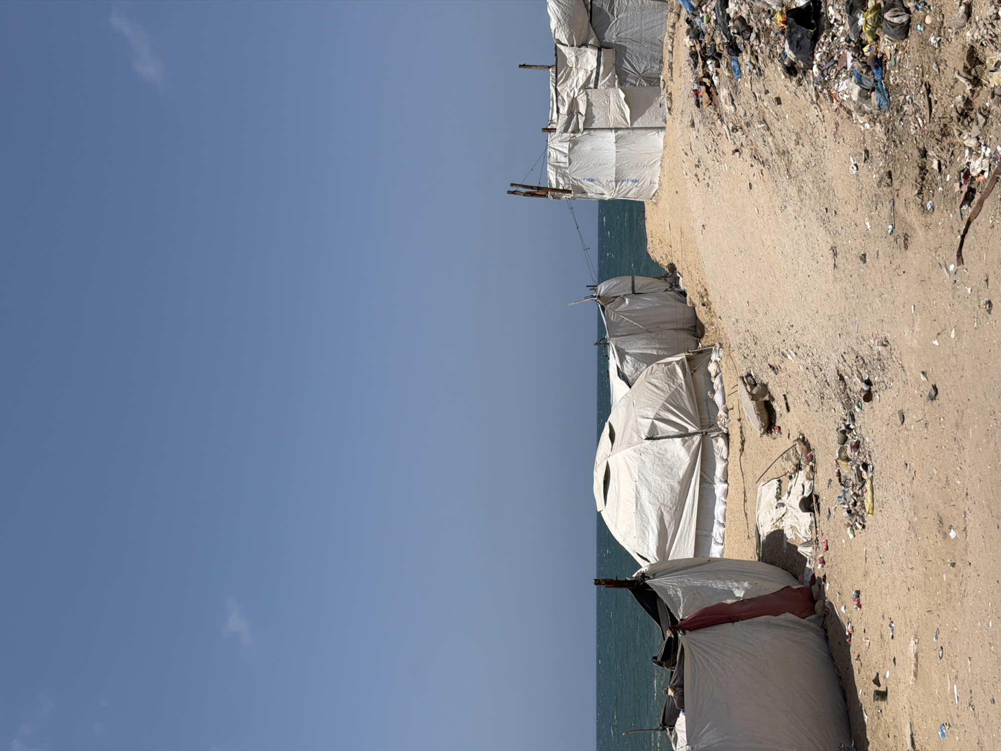 A low-angle, rotated outdoor shot depicts a coastal area, potentially a beach, with temporary shelters constructed along the shoreline. The shelters appear to be made of white tarpaulin or similar material, with wooden supports visible. A significant amount of debris and refuse litters the ground around the shelters. In the background, the blue ocean stretches to the horizon, meeting a clear, light-blue sky with a few small clouds. There are no people visible in the image. The location is Unknown, Unknown.