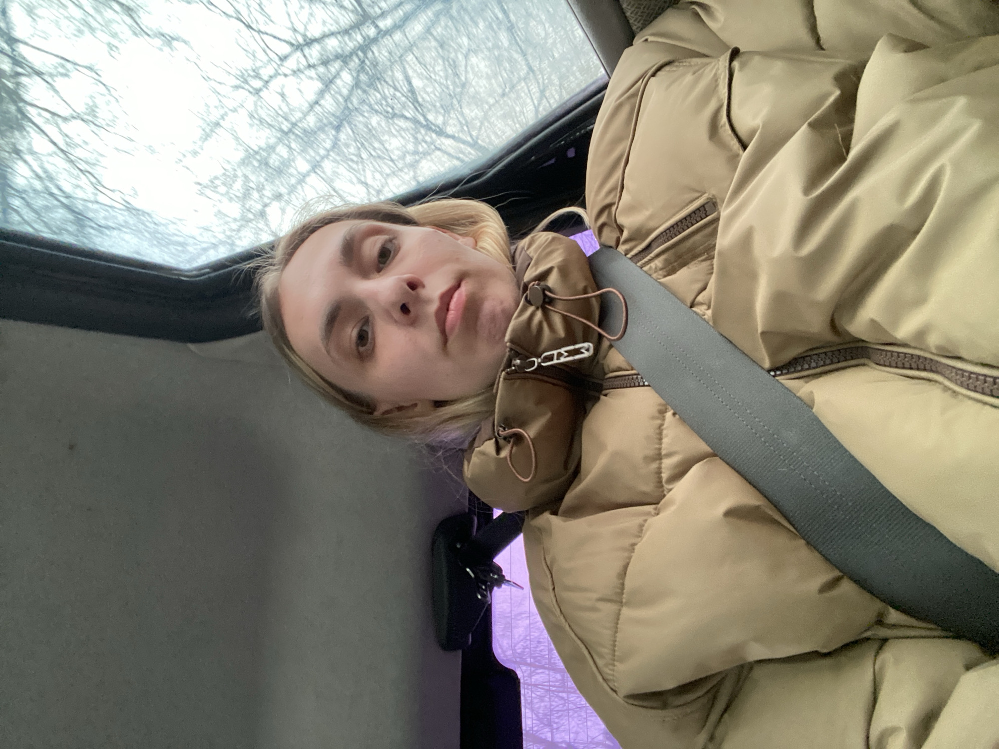 A woman with blond hair is sitting in a car, wearing a tan puffer jacket and a seatbelt. The view through the car window and sunroof shows trees and a gray sky. The location is Chaikovsky, Russia.