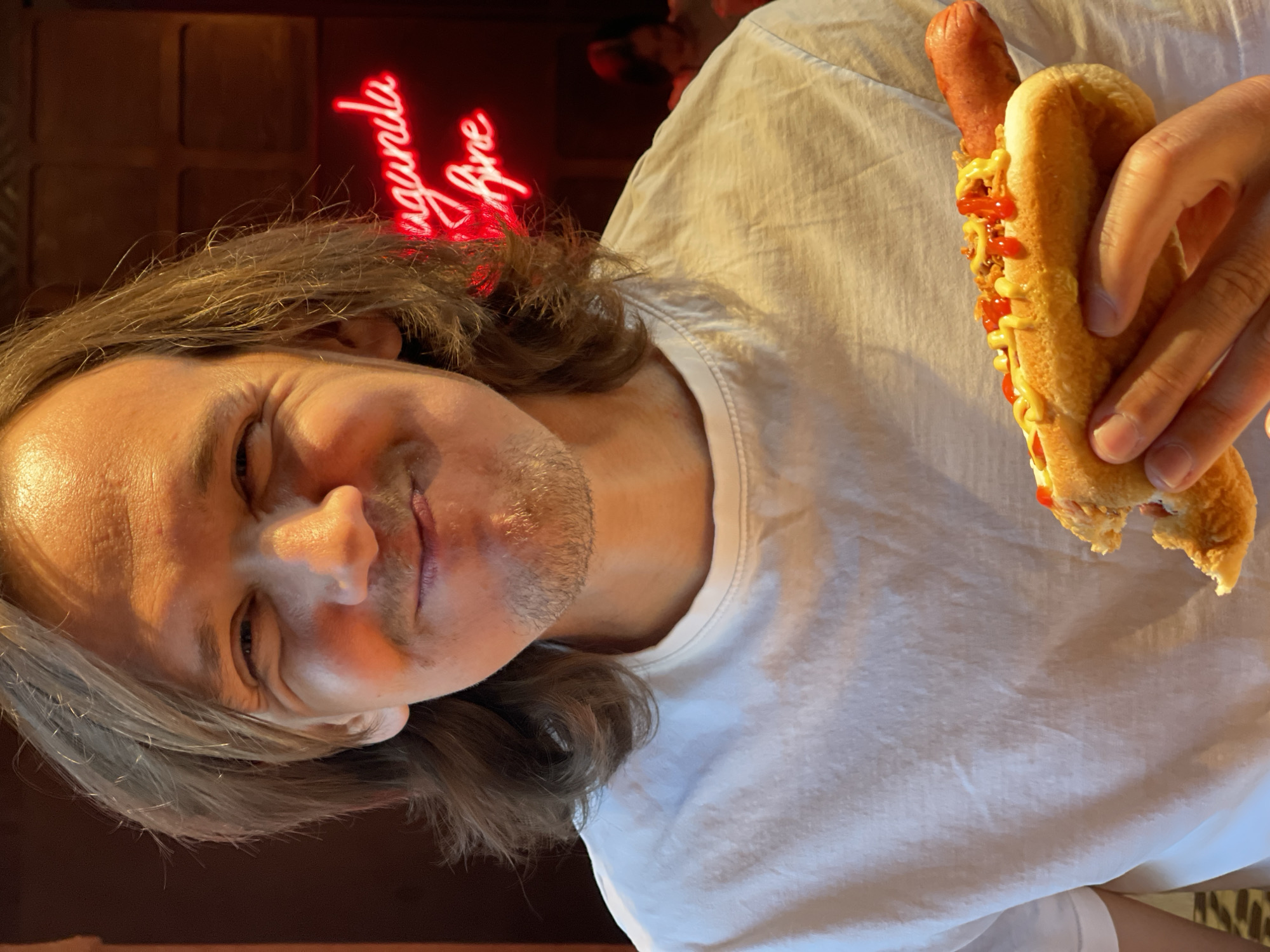 A man with long, light brown hair and a light beard is holding a partially eaten hot dog. The hot dog has mustard and ketchup. The man is wearing a white t-shirt. Behind him, there is a dark wooden wall with a red neon sign that says "ugunula Are." The location is Izhevsk, Russia.