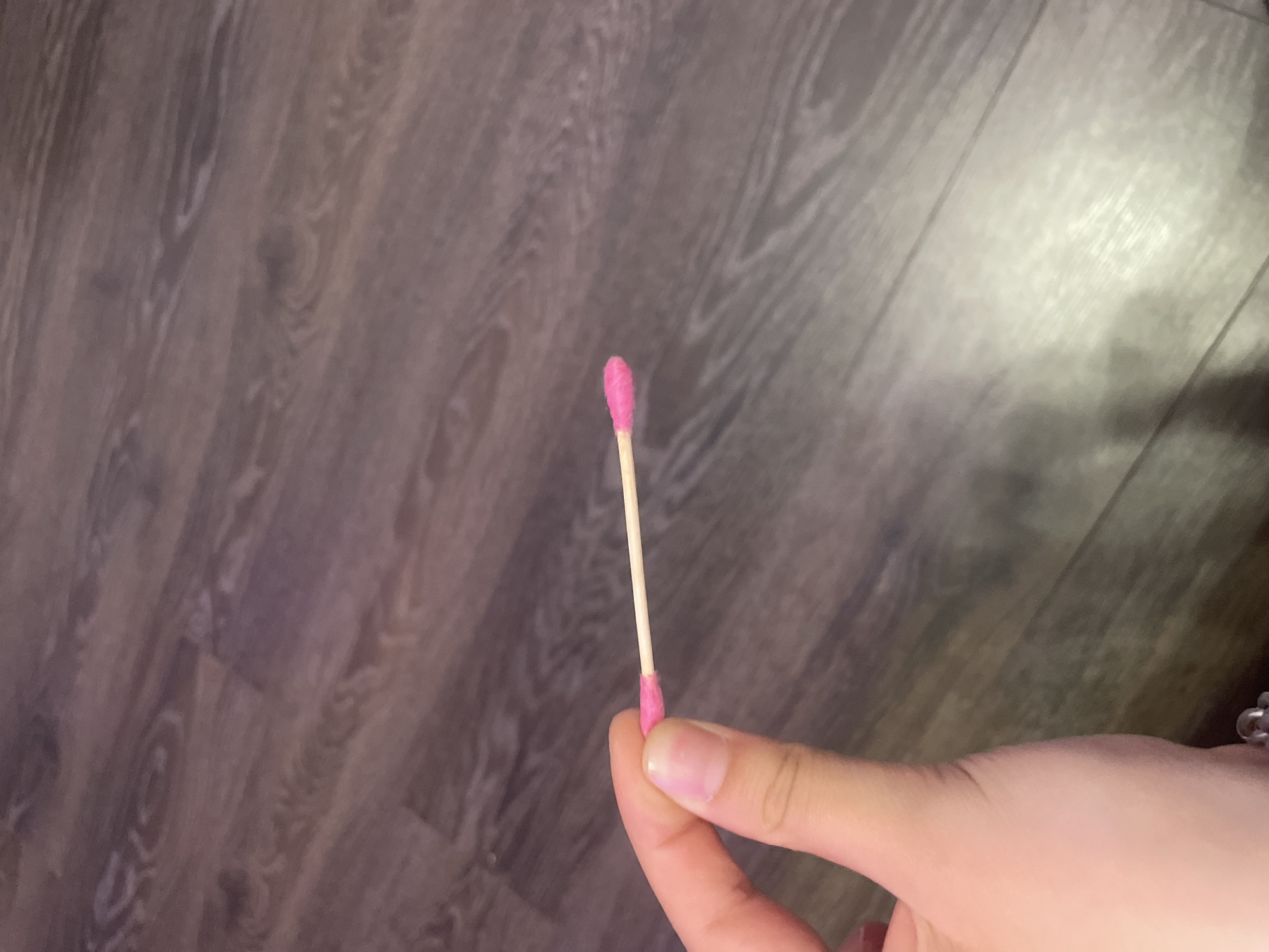 A person is holding a pink-tipped cotton swab horizontally with their fingers. The background is a wooden floor with a dark brown and grey grain pattern. The image is a close-up of the cotton swab and the hand holding it. There is no discernible location in the city of Antonovka, Russia.