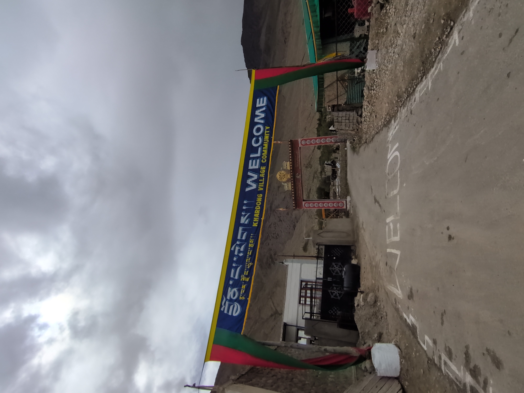 The image depicts an entrance archway to Khardung Village, India, with a large banner displaying "WELCOME" in English and Tibetan script. The banner is suspended across a road that leads into the village. The road surface has "WELCOME" painted on it in white capital letters. The archway itself is adorned with a decorative frieze and Buddhist prayer flags. To the left of the road, there is a building with a white facade and a metal gate. To the right, a green awning covers a structure. The background shows a mountainous, arid landscape under a cloudy sky. There are no people visible in the image.