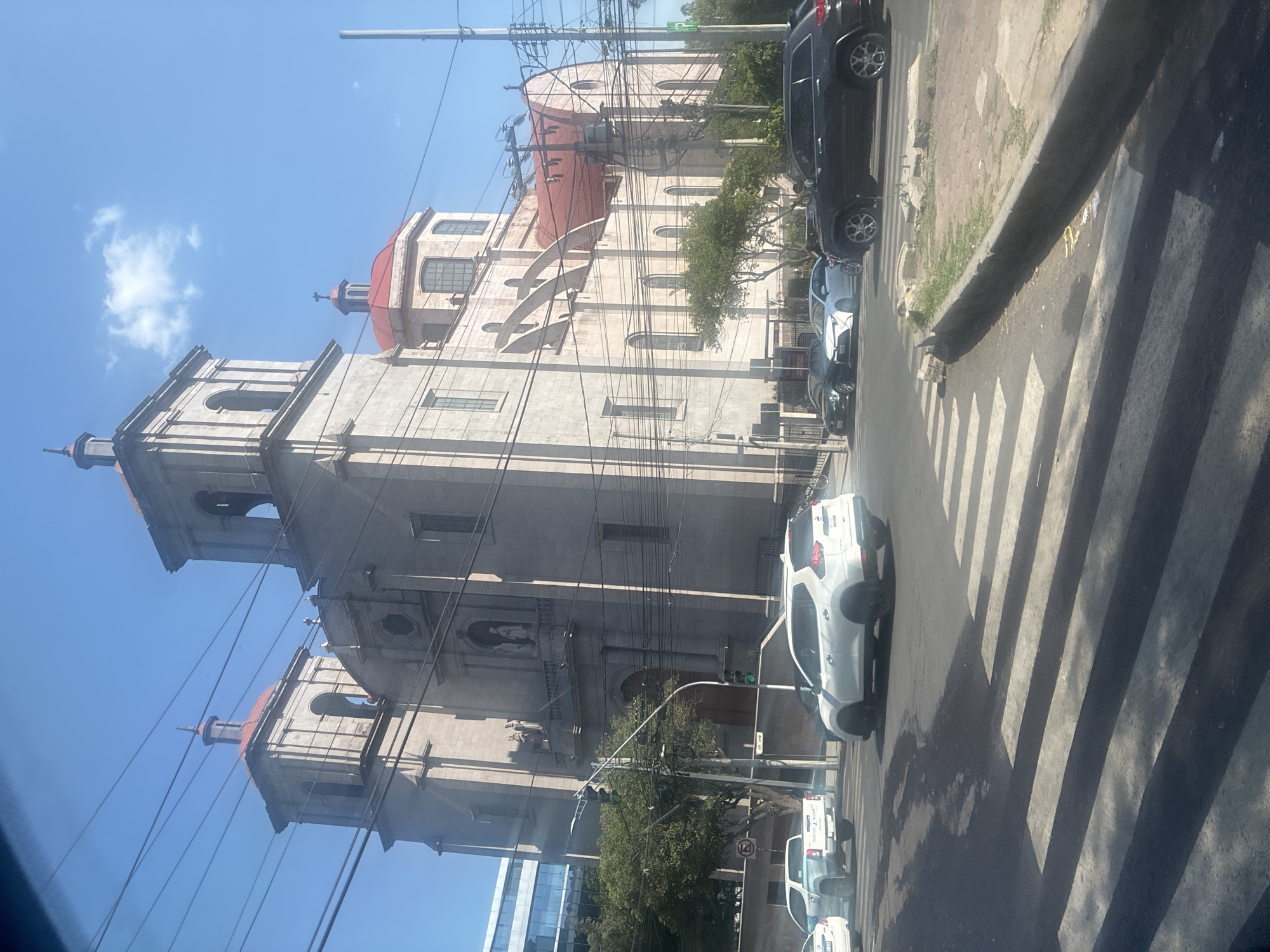 This media file captures a vibrant street scene in Mexico City, centered around a grand, light-colored stone building, likely a church or cathedral, characterized by its multiple red-domed roofs and two prominent bell towers with spires. The building's architecture features arched windows, decorative cornices, and classical elements.
The scene is set on a sunny day, indicated by the clear blue sky with scattered white clouds and the strong, distinct shadows cast on the street, suggesting midday or early afternoon. A paved street runs alongside the building, featuring a prominent zebra crossing in the foreground. Several vehicles are parked along the street; these include a dark SUV, a white SUV (possibly a Renault Duster), and further down, a light blue pickup truck with the partial text "Internet..." visible on its side, along with other cars.
Numerous black power lines are strung across the frame, connecting to utility poles and partially obstructing the view of the architectural details. Green trees line parts of the street, adding natural elements to the urban landscape. In the distance, a modern glass-fronted building can be glimpsed, reflecting the sky. A "No Entry" road sign is also visible among the parked vehicles. The image portrays a static, everyday urban setting with no specific activity taking place.