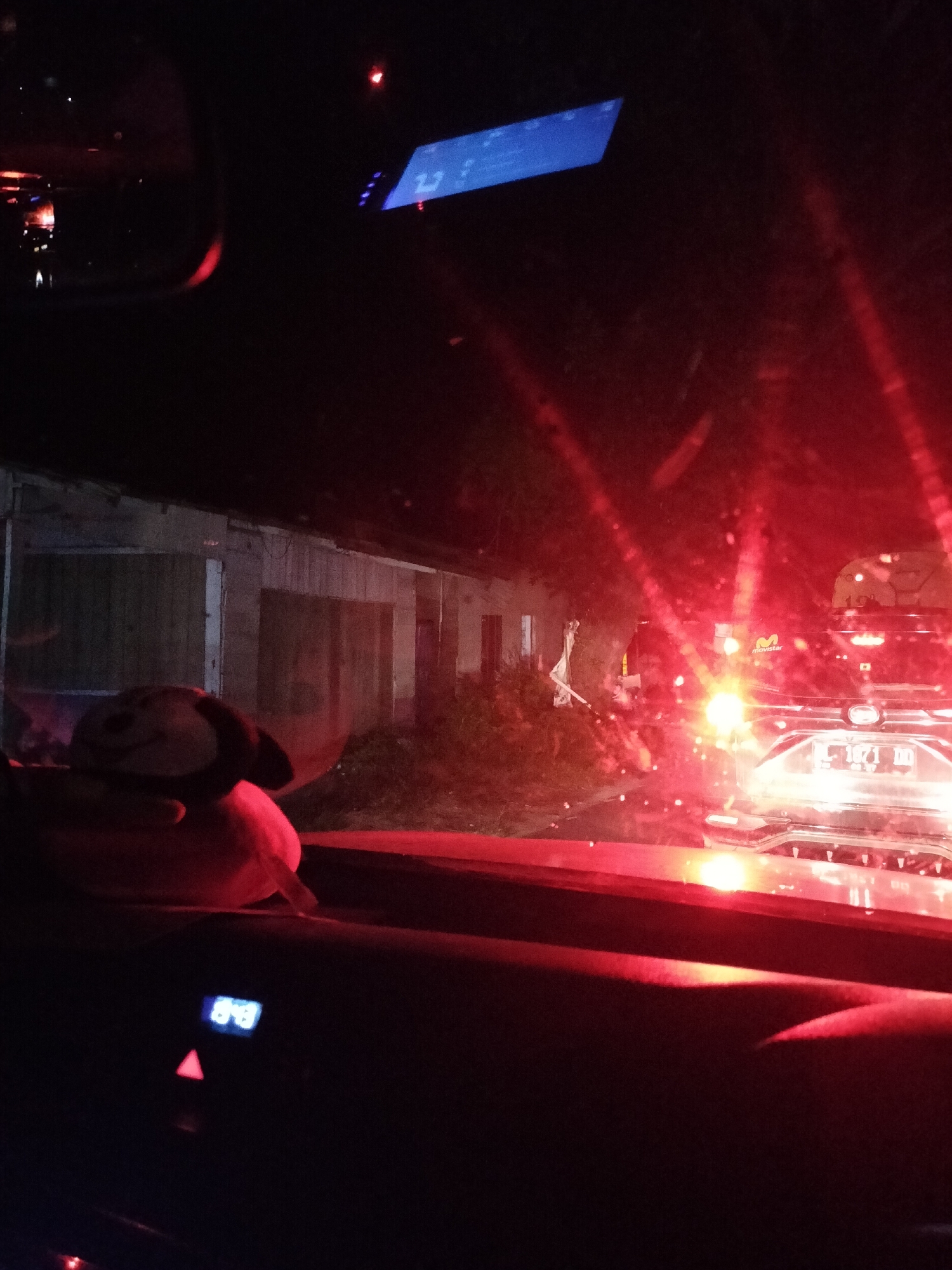 This image captures a nighttime view from inside a vehicle, likely in Alue Dua Bakaran Batee, Indonesia, judging by the context. The interior of the car is dimly lit, with a dashboard display showing "848" in blue numerals and a red triangular hazard light button. A stuffed toy, possibly a monkey with a smiling face, is visible on the dashboard. Outside the car, a street scene unfolds. Red taillights from other vehicles create a strong red glow and lens flares across the windshield. In the background, a building with a weathered wooden facade is visible, appearing dark and somewhat derelict. One of the vehicles ahead has a license plate that reads "L 1871 DD." The overall impression is one of being in traffic at night, possibly on a road with houses or businesses lining one side.