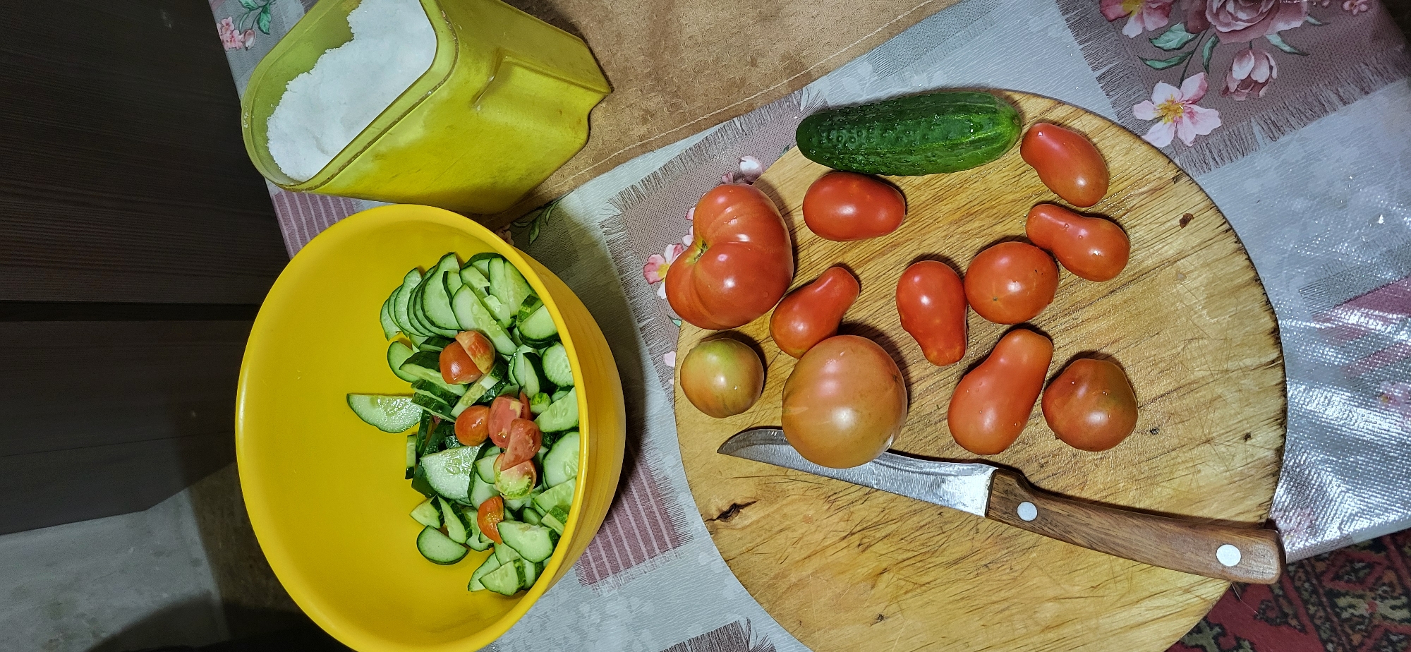 A rustic kitchen scene is depicted with a collection of fresh vegetables and ingredients arranged on a wooden cutting board and a yellow bowl. In the center of the image, a worn wooden cutting board displays a variety of tomatoes and a single cucumber. The tomatoes vary in size and shape, with some being plum-shaped and others round and larger. A knife with a wooden handle rests on the board, positioned next to a collection of tomatoes and a cucumber.
To the left of the cutting board, a bright yellow bowl is filled with sliced cucumbers and cherry tomatoes, suggesting a prepared salad. Beside the bowl, a lime-green container holds a generous amount of white salt. The scene is set on a table covered with a patterned tablecloth featuring floral designs in muted pinks and browns, with accents of silver thread. The background shows indistinct cabinetry and a wall, hinting at a domestic kitchen environment.
The image does not contain any discernible information that would allow for the identification of its location within the city of Oskemen, Kazakhstan. The scene is static and shows a still life arrangement of food preparation items. There are no people present, and no actions or interactions are depicted. The lighting appears to be from an overhead source, creating soft shadows. The overall impression is one of simple, home-style cooking.