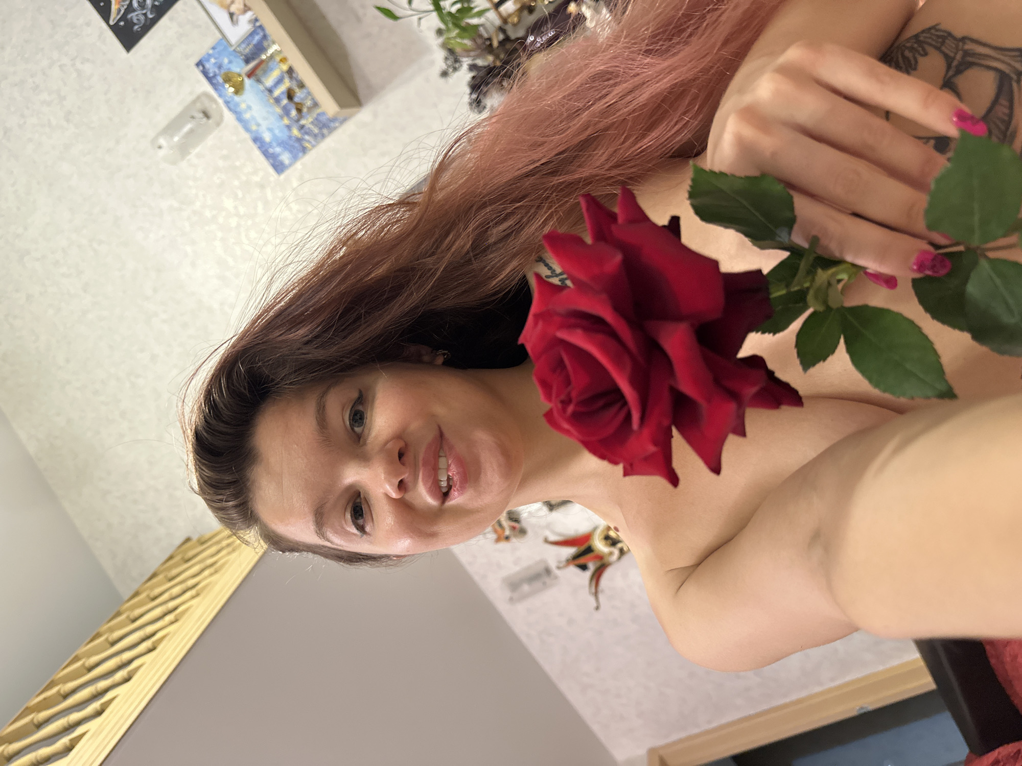 A woman with long, reddish-brown hair holds a red rose in her right hand, the stem extending downwards. She is looking towards the camera with a slight smile, her mouth open and biting her lower lip. Her bare shoulders are visible, and a tattoo is partially visible on her right upper arm. The background appears to be an indoor setting with textured wallpaper and shelves adorned with various decorative items, including what appear to be Venetian masks and framed pictures. The lighting suggests an indoor environment. There are no explicit indicators of the city of Vladimir, Russia, in the image or its context.