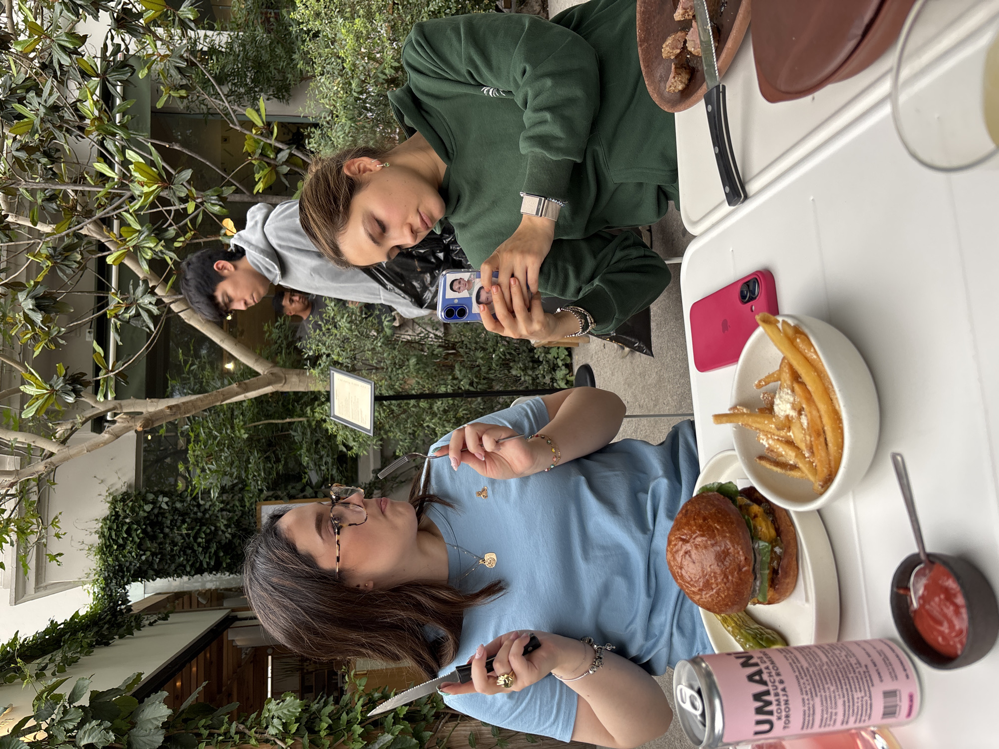 This daytime image captures a vibrant outdoor dining scene in what appears to be a garden or conservatory restaurant in Mexico City. Three individuals are seated around a white table, surrounded by lush green foliage.
In the foreground, a woman with dark hair and glasses, wearing a light blue t-shirt and multiple pieces of jewelry including bracelets, rings, and a necklace, holds a knife and fork. She is focused on her meal, which consists of a large burger with a dark bun, a pickle spear, and a small white bowl of french fries, some topped with a light seasoning. Beside her meal is a small black dish of ketchup and a pink can of "UMANI KOMBUCHA & TORONJA" (grapefruit kombucha), with additional text like "CON SABOR NATURAL Y JUGO DE FRUTA" visible.
To her left, another woman with light brown hair, wearing a dark green hoodie, is actively taking a photo of the food with her smartphone. Her phone, in a blue case, features two small photo inserts. She wears a watch and bracelets on her left wrist. On the table near her, a hot pink iPhone with a visible Apple logo rests next to a rustic brown plate holding pieces of meat and a serrated knife. A clear glass containing a light-colored beverage is also partially visible.
In the background, a young man in a grey hoodie is seen from the side, observing the activity. The setting is rich with natural elements, including trees, various green plants, and ivy-covered walls, suggesting an open-air or glass-enclosed dining area. A reflection of a person or scenery is visible in a glass surface behind the man. A white rectangular sign with black text, likely a menu, is partially visible hanging among the trees.