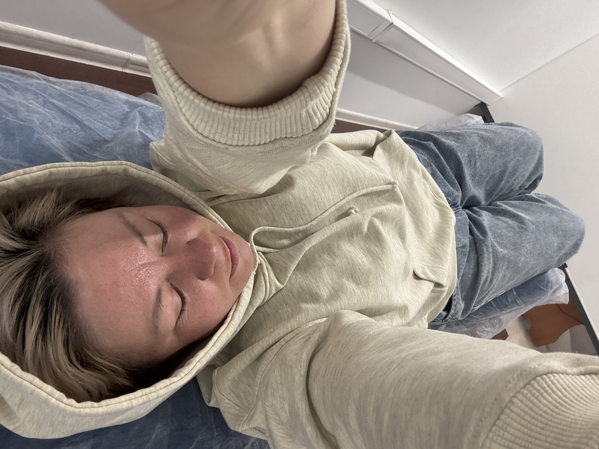 An overhead selfie captures a person with short, light-colored hair, dressed in a light beige hooded sweatshirt and faded blue denim jeans, lying down. The person's eyes are closed, and their face appears relaxed, showing a slight sheen on the forehead and cheek, which could be due to moisture or a skincare product. They are positioned on a crinkled, semi-transparent light blue sheet, which covers the surface beneath them. The background reveals a simple indoor setting: a plain white wall with a brown baseboard or trim running along its bottom edge, and a white ceiling from which a rectangular white light fixture is mounted. A dark, indistinct object, possibly a furniture support or bracket, is faintly visible near the ceiling. The perspective of the image indicates it was taken by the person themselves, with their extended arm and hand holding the camera visible at the top of the frame. The presence of the blue sheet and the clinical yet neat environment might suggest a professional setting such as a spa, salon, or medical office in Chaikovsky, Russia. No text is visible in the image.