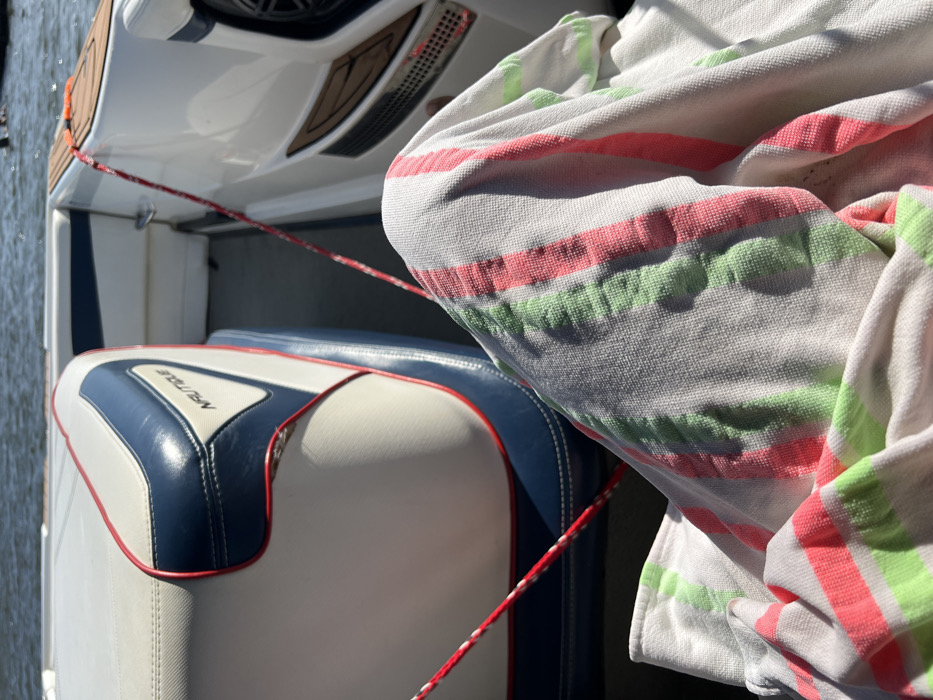 A first-person perspective image depicts the interior of a recreational powerboat on water in Coruche, Portugal, under bright daylight. In the foreground, the upper legs of an individual are partially visible, covered by a white terry cloth towel featuring prominent vertical stripes in neon green and coral pink. To the left, a white marine seat with dark blue sections and red piping is present, displaying a "NAUTIQUE" brand logo on its side. To the right, another section of the boat's white interior hull features dark brown textured marine flooring and a circular speaker grill. A red and white braided rope extends diagonally across the boat's floor from the upper left to the lower right of the frame. The background shows a body of water with a gently rippled surface, suggesting the boat is afloat on a river or lake. A blurred, dark shape is faintly visible on the water in the distant background.