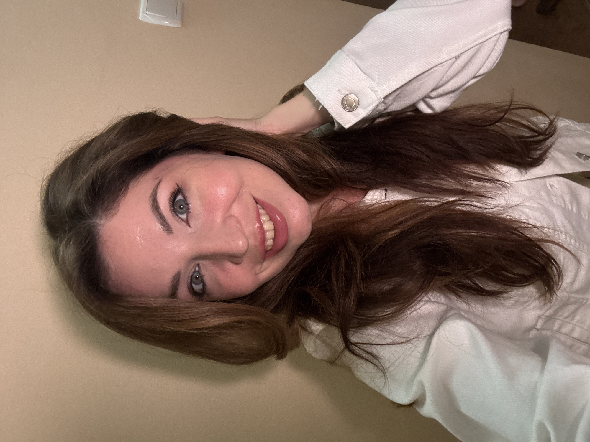 A young woman with long, brown hair and blue eyes smiles into the camera. She is wearing a white denim jacket and her right hand is resting on her head, pushing her hair back. The background is a plain, beige wall with a white light switch visible on the right side of the frame. The lighting suggests an indoor setting, likely a home or hotel room, with a warm tone. There is no identifiable information about the location in the city of Odesa, Ukraine.