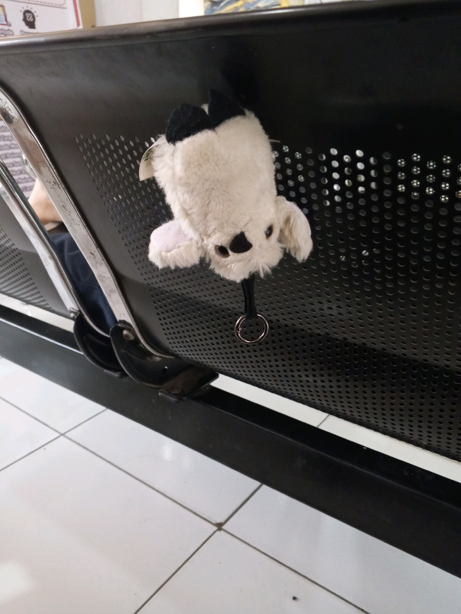 A fluffy, white stuffed animal with black accents hangs from a metal ring attached to a perforated black bench. The bench appears to be in a public waiting area, possibly at a transportation hub or clinic, given the tiled floor and the presence of other similar benches. The lighting suggests an indoor environment. No specific location in Karawang, Indonesia can be identified from the image alone.